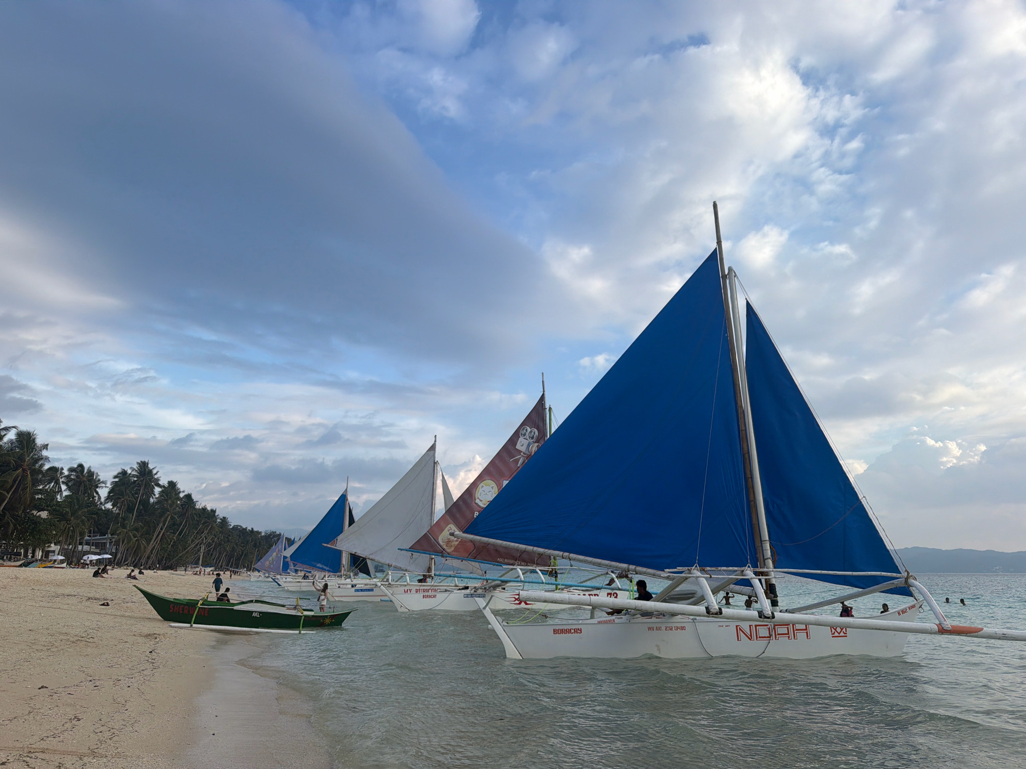 A group of sailboats with blue and white sails are moored in the shallow waters off a sandy beach. The beach is lined with palm trees and a few scattered beachgoers. The sky is cloudy with patches of blue. In the foreground, a green sailboat with "SHERWINE AKL" written on its side is partially beached. Behind it, several larger sailboats are visible, some with passengers. The sailboat in the most prominent position in the foreground has "NOAH" written in large red letters on its hull. The scene appears to be of Boracay Island, a tourist destination in Malay, Aklan, Philippines, known for its white sand beaches and water activities.