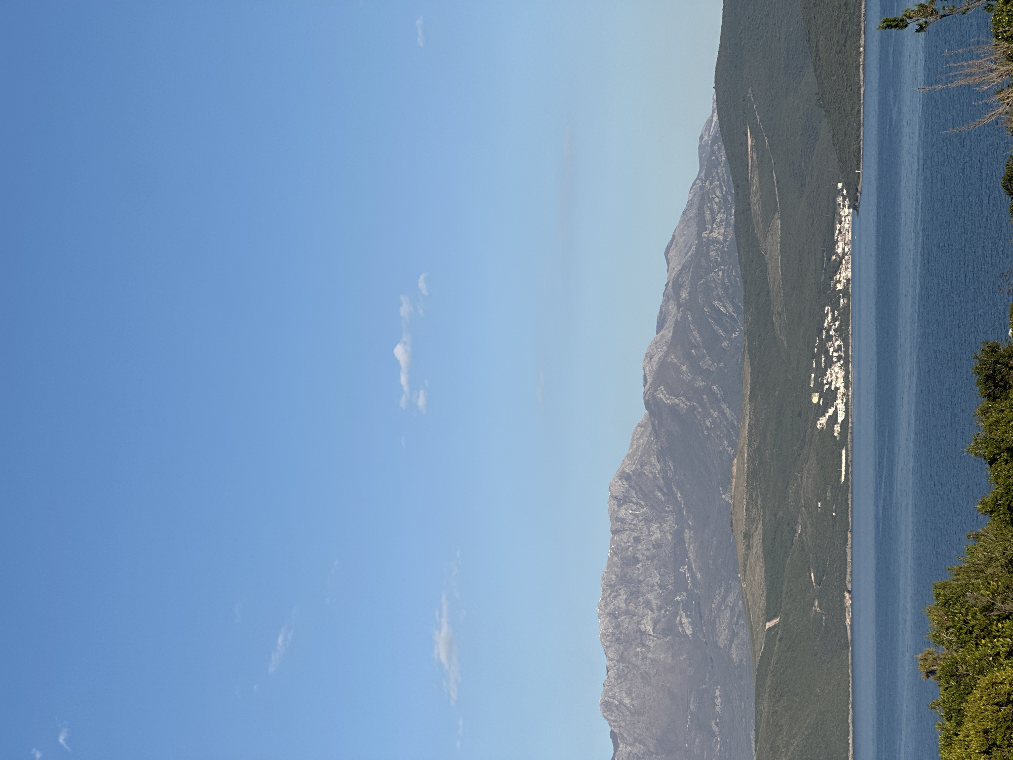 The image depicts a coastal landscape in Glavaticici, Montenegro. On the right is a narrow strip of land where the town resides between a mountain range and the sea. The town appears as a line of predominantly white buildings hugging the coastline. A mountain with green vegetation rises steeply behind the town. To the left of the mountain range is a vast blue sky with a few scattered clouds. The sea is a deep blue color and extends into the distance. Some trees are visible on the right edge of the frame.
