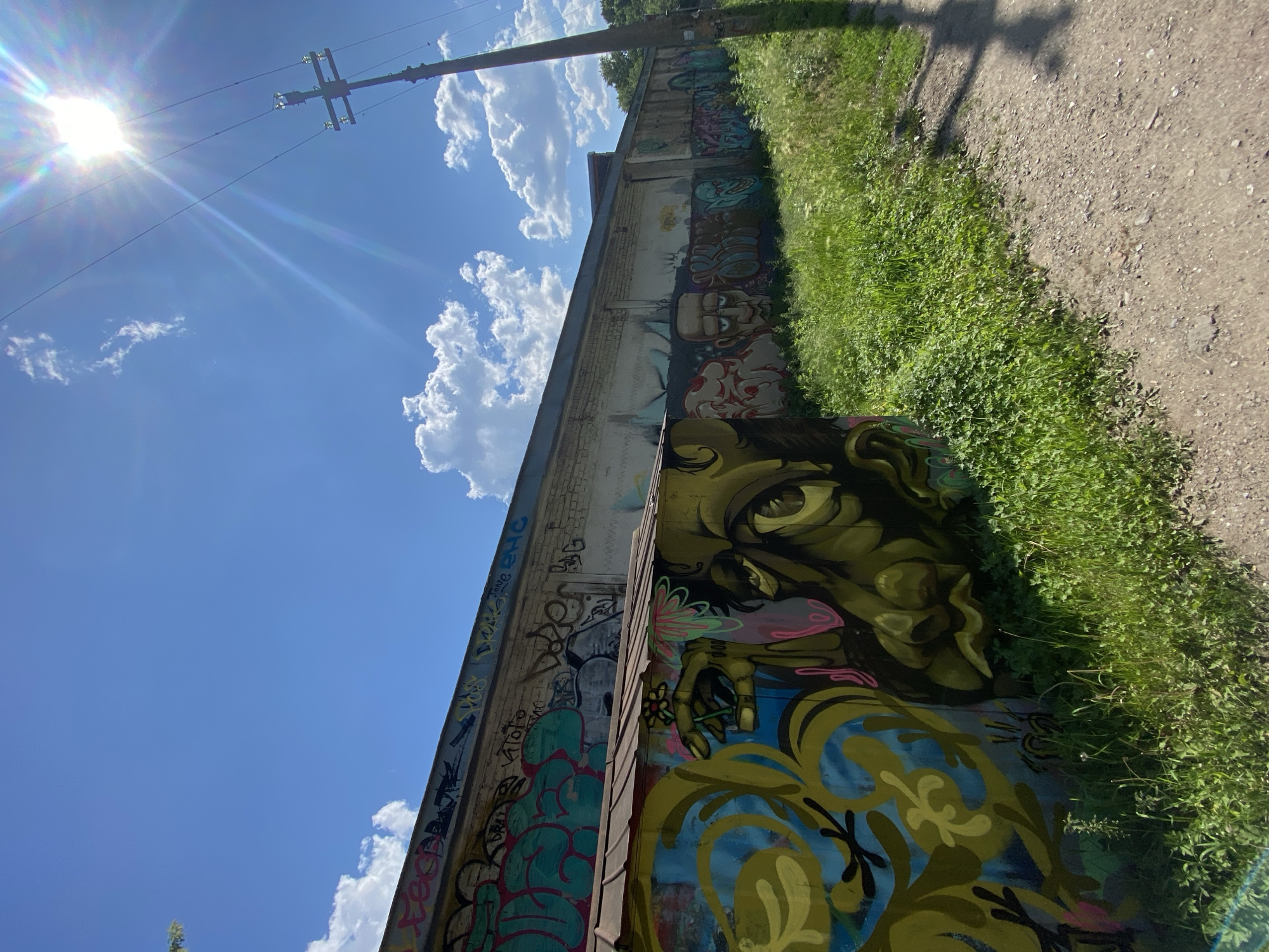 The image shows a long, cream-colored building covered in graffiti, stretching from left to right. The building has a metal roof on a smaller structure on the left. Lush green grass and weeds grow along the base of the building and a dirt path runs parallel to it on the right. A utility pole with wires stands to the right of the building, silhouetted against a bright, sunlit blue sky with scattered white clouds. The sun is high and glinting, creating lens flare. The graffiti on the building is colorful and varied, featuring large abstract shapes, lettering, and stylized faces. The overall impression is one of urban decay and artistic expression.
This scene is located in Khabarovsk, Russia, specifically at the abandoned industrial complex in the Zheleznodorozhny District. The complex, which was once a large factory, is now a canvas for local graffiti artists, transforming the decaying structures into a vibrant outdoor gallery. The area is known for its unique blend of industrial remnants and street art, attracting photographers and urban explorers alike.