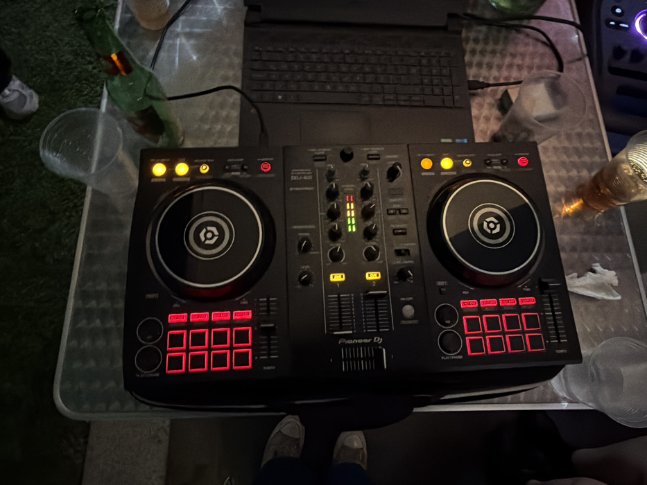 This nighttime image, captured in San Bernardino Tlaxcalancingo, Mexico, depicts an outdoor DJ setup on a metallic-surfaced table. A black Pioneer DDJ-400 DJ controller with active red and yellow indicator lights is centrally positioned. To its right, a black laptop is connected and open. The table surface exhibits a textured, possibly corrugated pattern, with reflections from an unseen light source. Several clear plastic cups are scattered across the table. A green glass beer bottle, partially full, is visible near the top right, and another clear glass bottle containing an amber liquid is situated towards the bottom. A small crumpled white tissue also rests on the table. In the immediate background, dark green grass is discernible, suggesting a lawn or garden setting. The lower halves of two pairs of feet, shod in sneakers, are visible on the left side, indicating the presence of at least two individuals standing near the setup. The overall scene is dimly lit.
