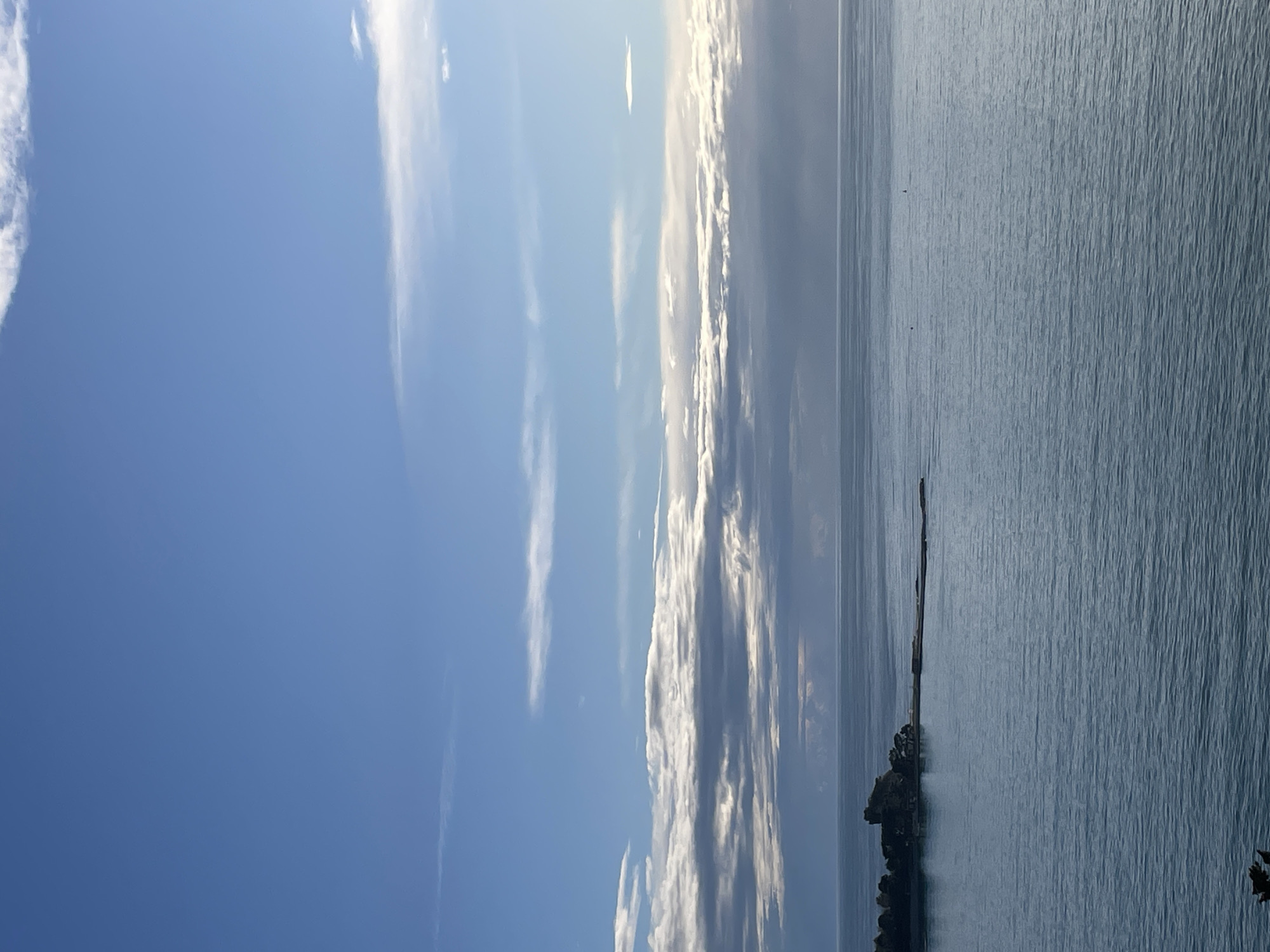 The image displays a seascape captured in Budva, Montenegro, looking towards Sveti Nikola Island. The scene is dominated by a blue sky with horizontal bands of white and grey stratocumulus clouds, thicker and darker towards the horizon line on the right, and thinner, scattered cirrus-like clouds higher up. The upper portion of the sky is clear blue.
The middle and lower sections of the frame show the calm, rippled surface of the sea, appearing blue-grey under the sky's light. On the left side, Sveti Nikola Island is visible, characterized by dark, rocky formations covered with dense, dark green vegetation, consistent with trees or shrubs. A long, narrow, dark grey landmass or causeway extends from the island's eastern side into the sea, stretching towards the center-right of the frame. Two small, dark, cylindrical buoys are present in the open water in the mid-distance, positioned towards the right of the frame. No individuals, animals, or active interactions are visible. The overall lighting suggests daylight conditions.