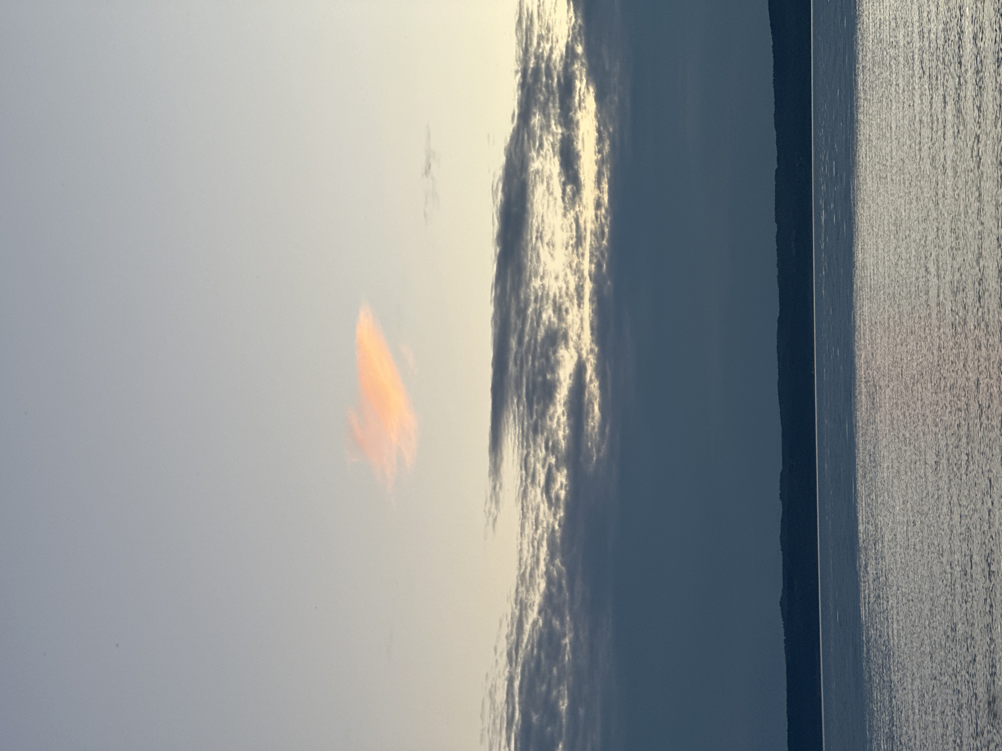 The image shows a serene body of water with a distant, dark treeline under a twilight sky. The sky transitions from a pale blue at the top to a soft yellow and then a deep blue closer to the horizon. A few wispy clouds are visible, with one small cloud in the upper portion of the frame tinged with a warm orange hue, possibly from the setting or rising sun. The water is calm, with gentle ripples reflecting the light from the sky. The treeline is a dark, silhouetted band across the horizon. The scene appears to be a natural landscape, likely a lake or a large river, given the calm water and the distant treeline suggesting a vast expanse of nature rather than an urban environment. No people or man-made structures are visible, contributing to the tranquil and remote atmosphere of the scene.