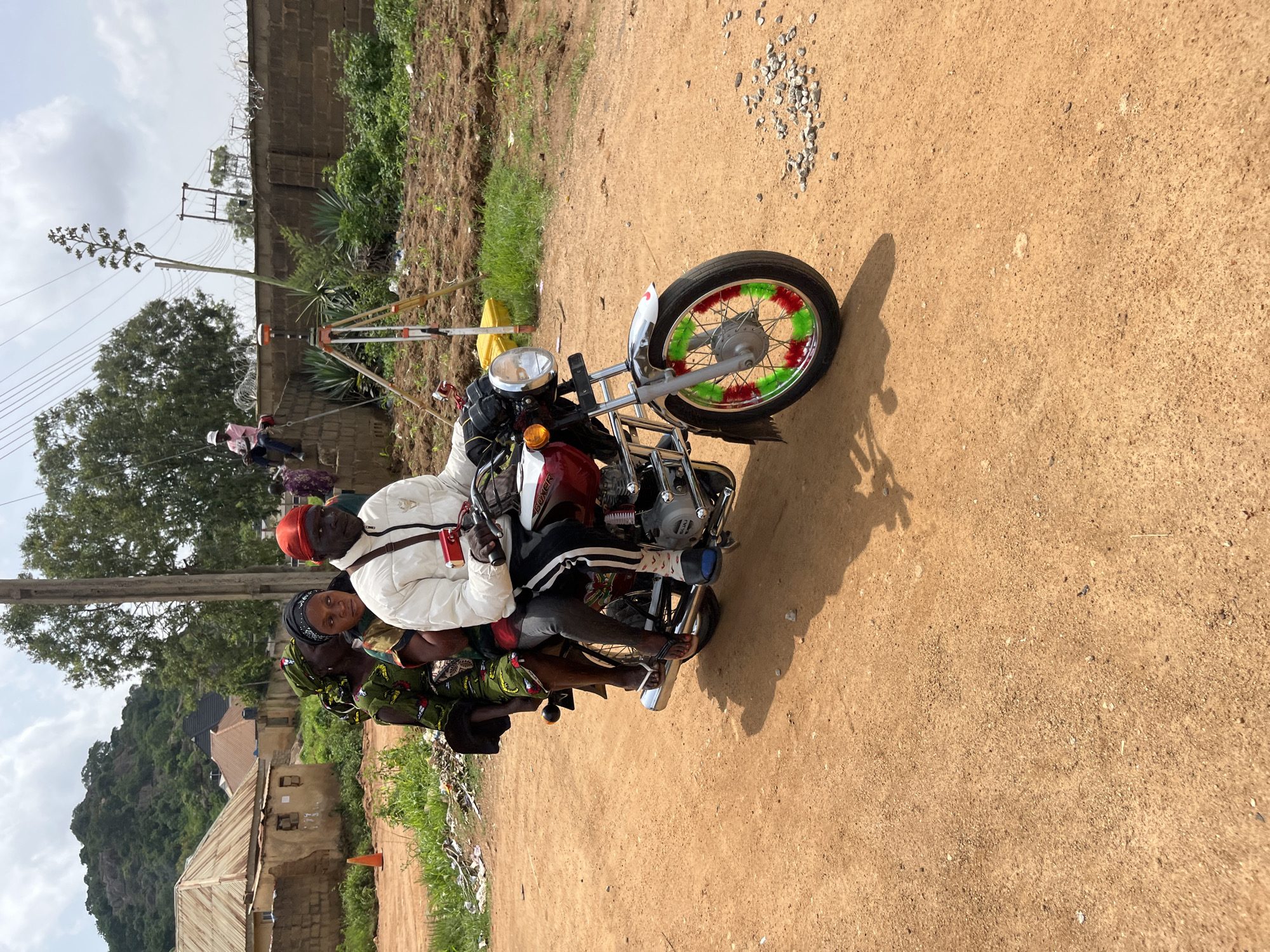 In Bukuru, Nigeria, three people are riding on a motorcycle. The driver, a man wearing a red cap and a white jacket, is in the front. Behind him, two women are seated, one in the middle and another in the back, both wearing headscarves. The motorcycle is a red and white Boxer model. The scene takes place on a dirt road, with buildings and a hill visible in the background. In the background, there are also construction materials and survey equipment. The sky is cloudy.