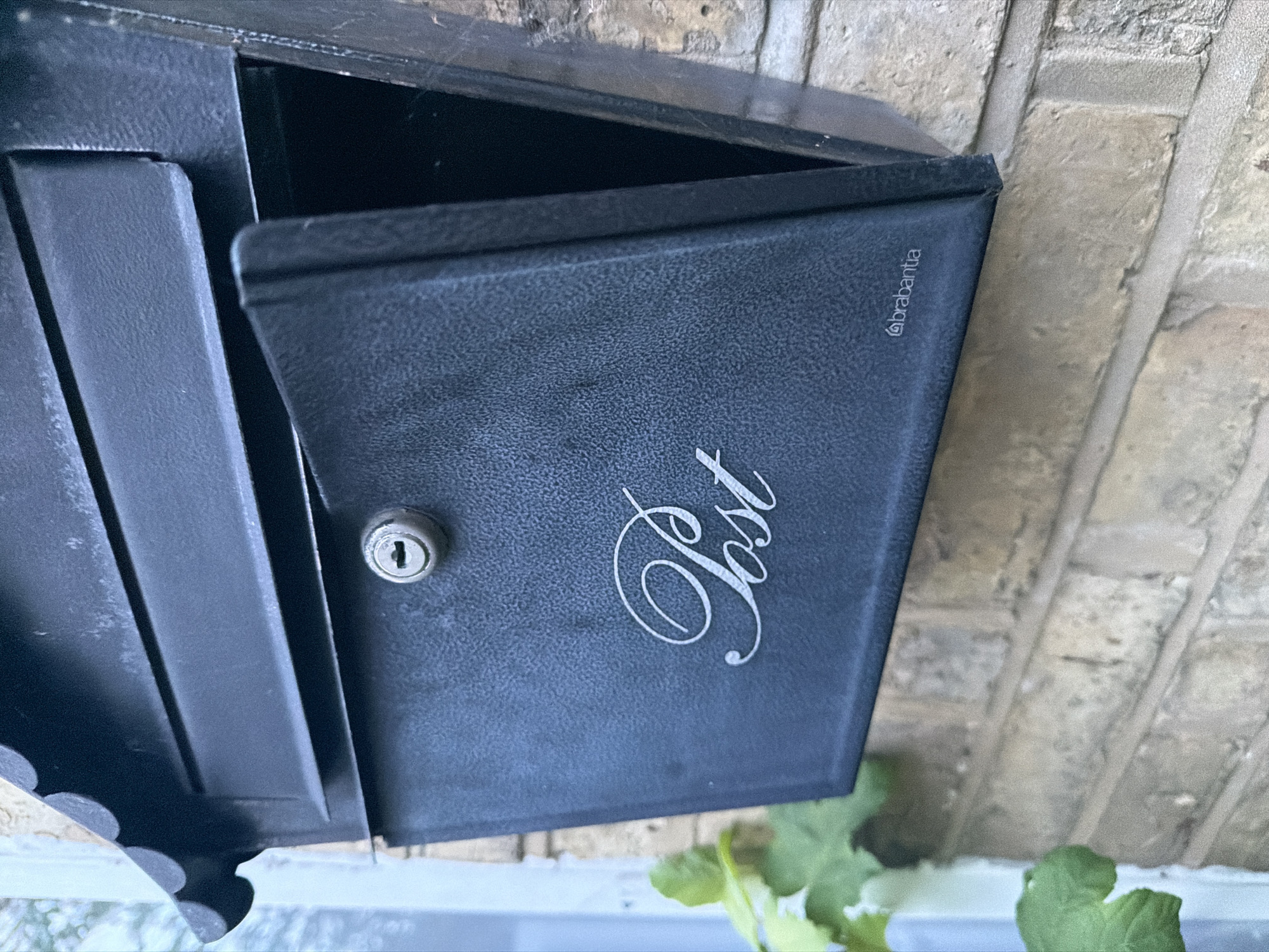 A dark grey, textured metal mailbox with a decorative, scallop-edged top cover is prominently displayed, mounted on a light brown brick wall. The front-access door of the mailbox is hinged at the bottom and stands open, revealing a dark interior. Centered near the top of the open door is a silver-colored keyhole. Below the keyhole, the word "Post" is inscribed in a decorative, calligraphic white script. In the bottom right corner of the door, the small "brabantia" logo is visible in white. To the left of the mailbox, green leaves from a plant are partially visible. A light-colored, vertical structural element, possibly a window frame, is present in the blurred background to the far left. The image does not contain sufficient identifying information to pinpoint its exact location within Maidstone, United Kingdom.