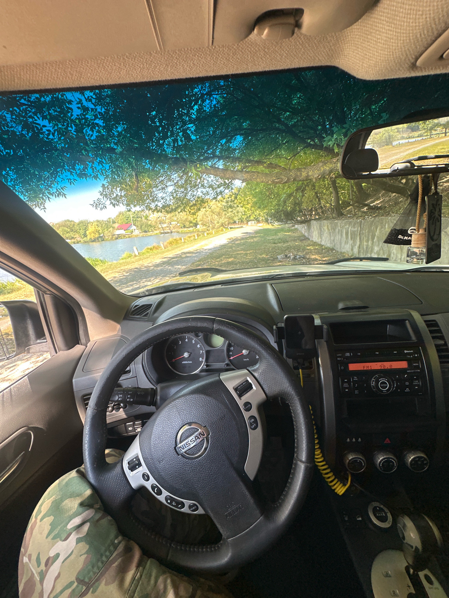 The image shows the interior of a car, looking forward through the windshield. The steering wheel, dashboard with gauges, and car radio are visible. Outside the car, a tree casts shadows on the windshield, and a body of water and a road are visible in the distance. A single person's leg, wearing camouflage pants, is visible on the left side of the frame. This image was taken in the city of Makiv, Ukraine.