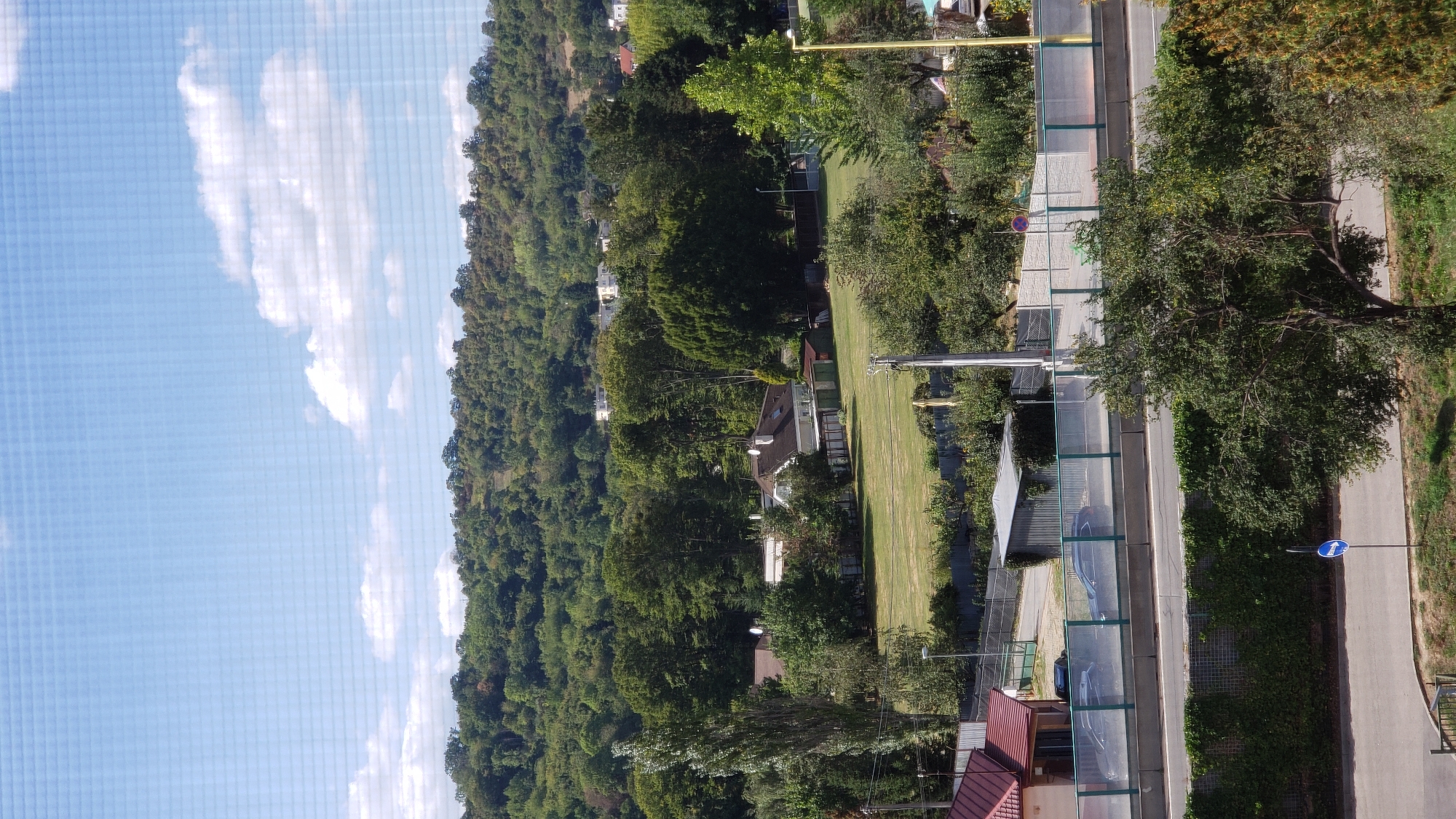The image depicts a suburban or semi-rural landscape with a densely wooded hill in the background. In the mid-ground, there is a large, open grassy field, possibly a park or sports field, with several houses and smaller structures surrounding it. A tall, slender tree stands prominently on the left side of the field. A road with a barrier and parked cars runs horizontally across the lower portion of the image, separating the field area from the foreground. The foreground shows more vegetation, including trees and bushes, and a small traffic sign indicating a directional route. The sky is blue with scattered white clouds. The scene appears to be taken from an elevated position, possibly from a window overlooking the area. The location is identified as Prešov, Slovakia, implying a view of the surrounding greenery and residential areas of the city.
