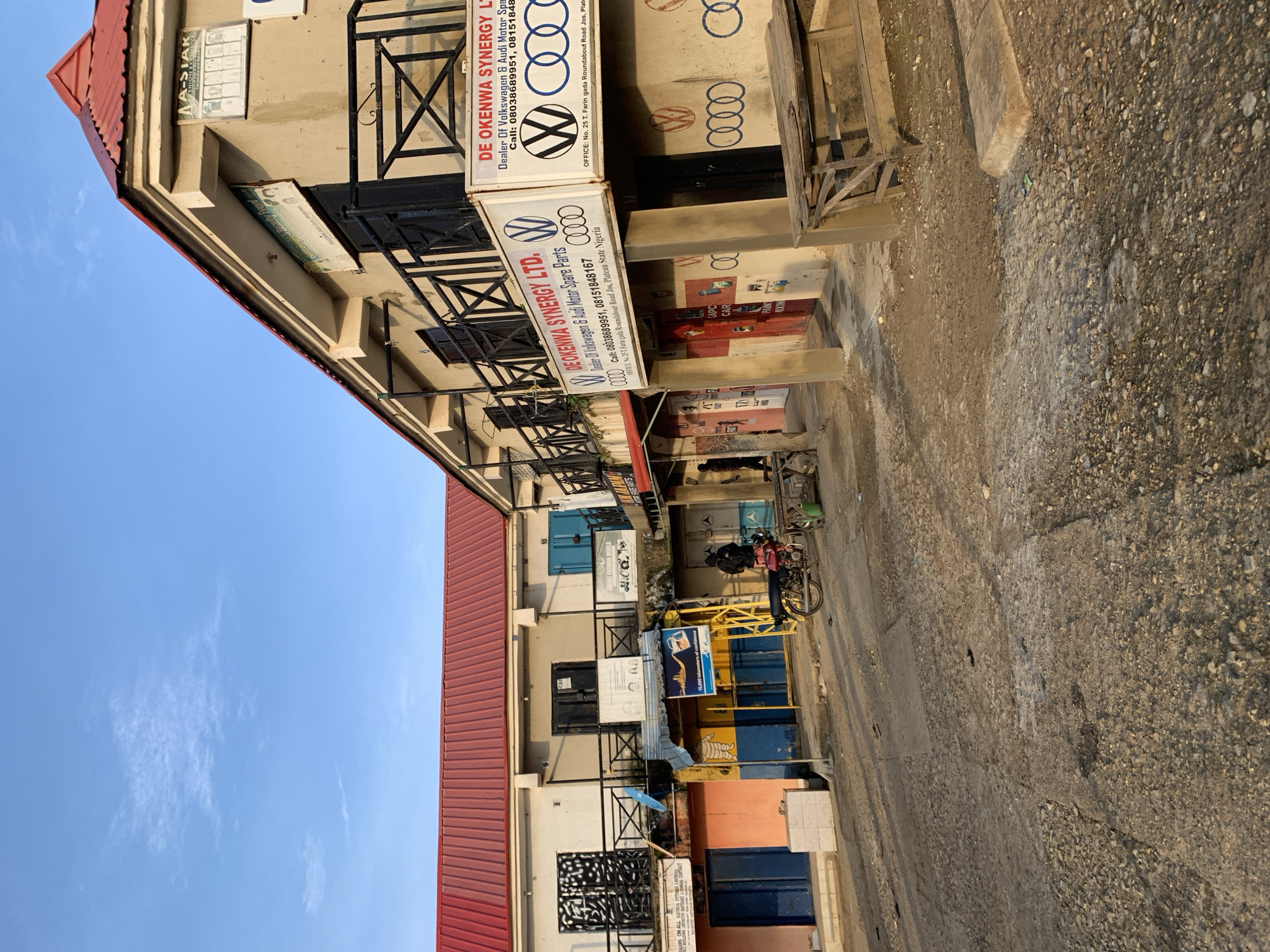 This is a frontal view of a street scene in Rafin Par, Nigeria. The image captures a row of commercial buildings with shops on the ground floor and residences or offices on the upper floors. The buildings have red corrugated metal roofs. On the right side of the image, a sign reads "DE OKENWA SYNERGY LTD. Dealer Of Volkswagen & Audi Motor Spare Parts". Below this, there are logos for Volkswagen and Audi. The street in front of the shops is paved with concrete and gravel, with some debris scattered on the ground. A motorcycle is parked on the right side of the street, next to a wooden bench. A person is visible standing near the motorcycle. The sky is blue with a few wispy clouds. The overall scene depicts a commercial area in a Nigerian town, likely during daylight hours.