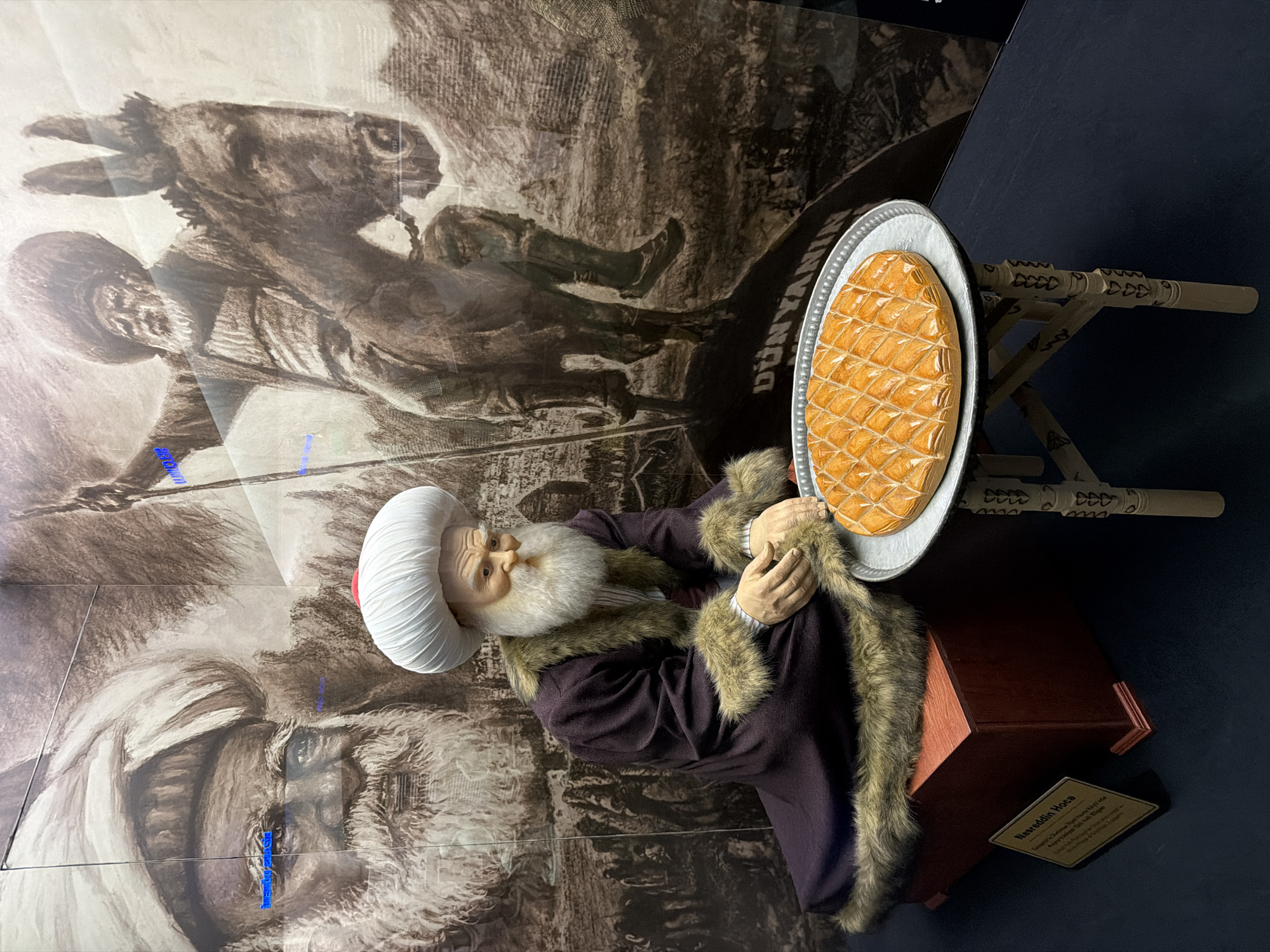 This vertical indoor image displays a museum exhibit featuring a life-size mannequin of Nasreddin Hoca, a renowned figure in Turkish folklore. The mannequin portrays an old man with a long, flowing white beard and a serene, wise expression. He is adorned in traditional attire, including a distinctive white pleated turban with a red cap, a dark purple robe, and a matching fur-trimmed cloak. He is seated on a dark wooden box, appearing to reach towards a large, golden-brown dessert, likely baklava or a similar pastry, arranged in a diamond pattern on a silver platter. The platter rests on a small, intricately carved wooden stand.
The background features a large, sepia-toned mural or artwork, visible through a reflective surface. This artwork depicts scenes related to Nasreddin Hoca, including an illustration of a man (presumably Hoca) riding a donkey, and a detailed close-up portrait of an old man's face. Partially visible text on the mural includes "DÜNYANIN" (of the world).
At the base of the exhibit, a small gold-colored plaque clearly identifies the figure as "NASREDDİN HOCA." Further text on the plaque, though partially obscured, includes references to "Ladik, Eskişehir," grounding the exhibit within the local context of Eskisehir, Turkey. The display effectively combines historical representation with a symbolic culinary element, likely a nod to Turkish culture. The scene is illuminated by indoor artificial lighting, which creates minor reflections on the protective surface covering the background mural.