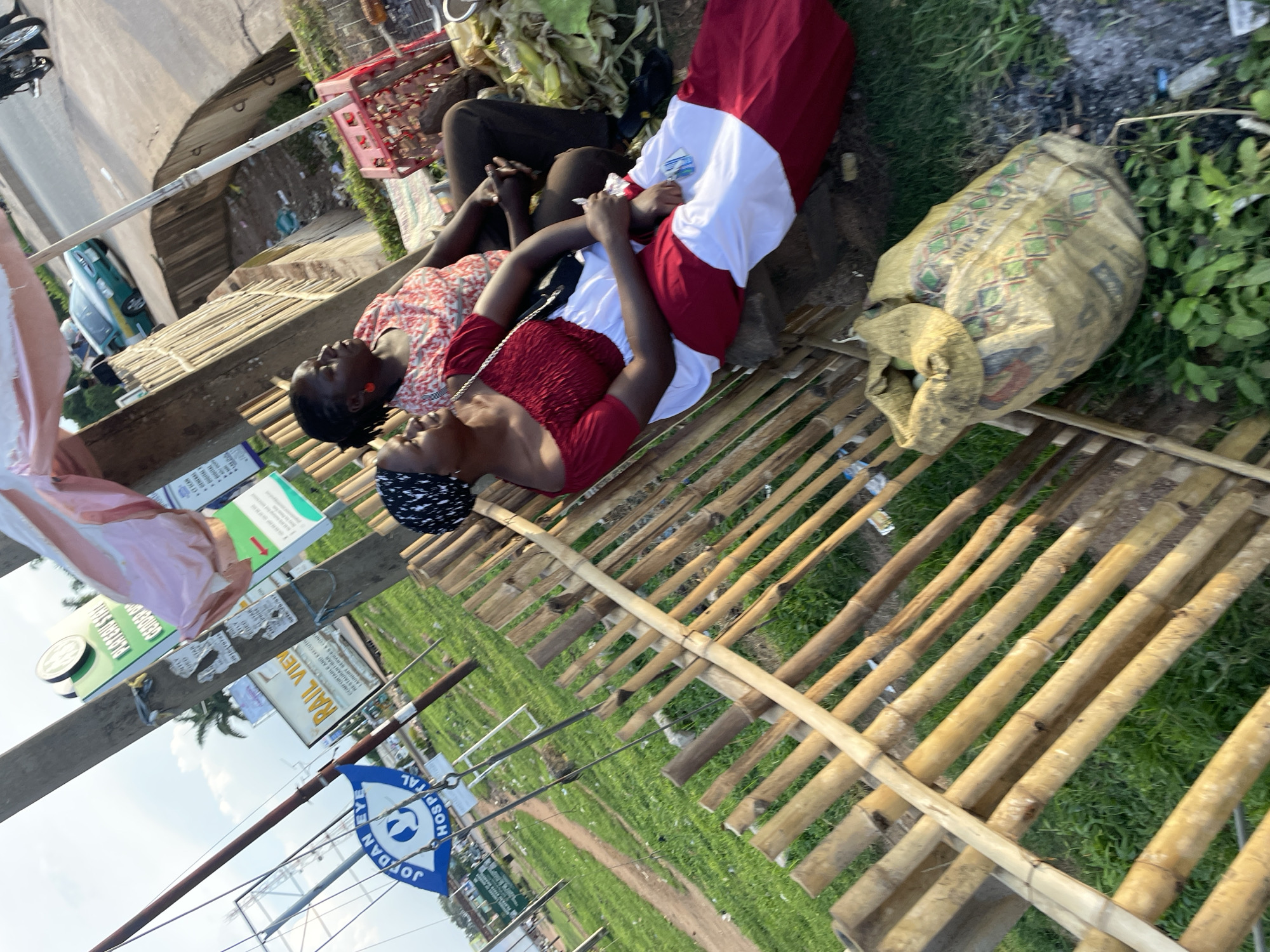 The image captures an outdoor scene, possibly along a roadside in Jos, Nigeria, under a bright, overcast sky. Two women are seated on a wooden bench, partially obscured by a bamboo fence and a light-colored canopy. The woman in the foreground wears a red and white off-the-shoulder dress and a black and white headscarf. She is looking to her right, with her hands resting on her lap. The second woman, behind and to the right of the first, is wearing a red and white patterned top and dark trousers. She is looking forward and has her hands clasped. In the background, several signs are visible, including one for "Jordan Eye Hospital." A green and white car is driving on a paved road in the distance, and a red plastic crate is placed near the women. The ground is covered with green grass and scattered debris, including a large, yellow woven bag filled with produce. A motorcycle is also partially visible in the upper right corner of the frame. The overall impression is of a casual, everyday moment captured in a bustling urban environment.