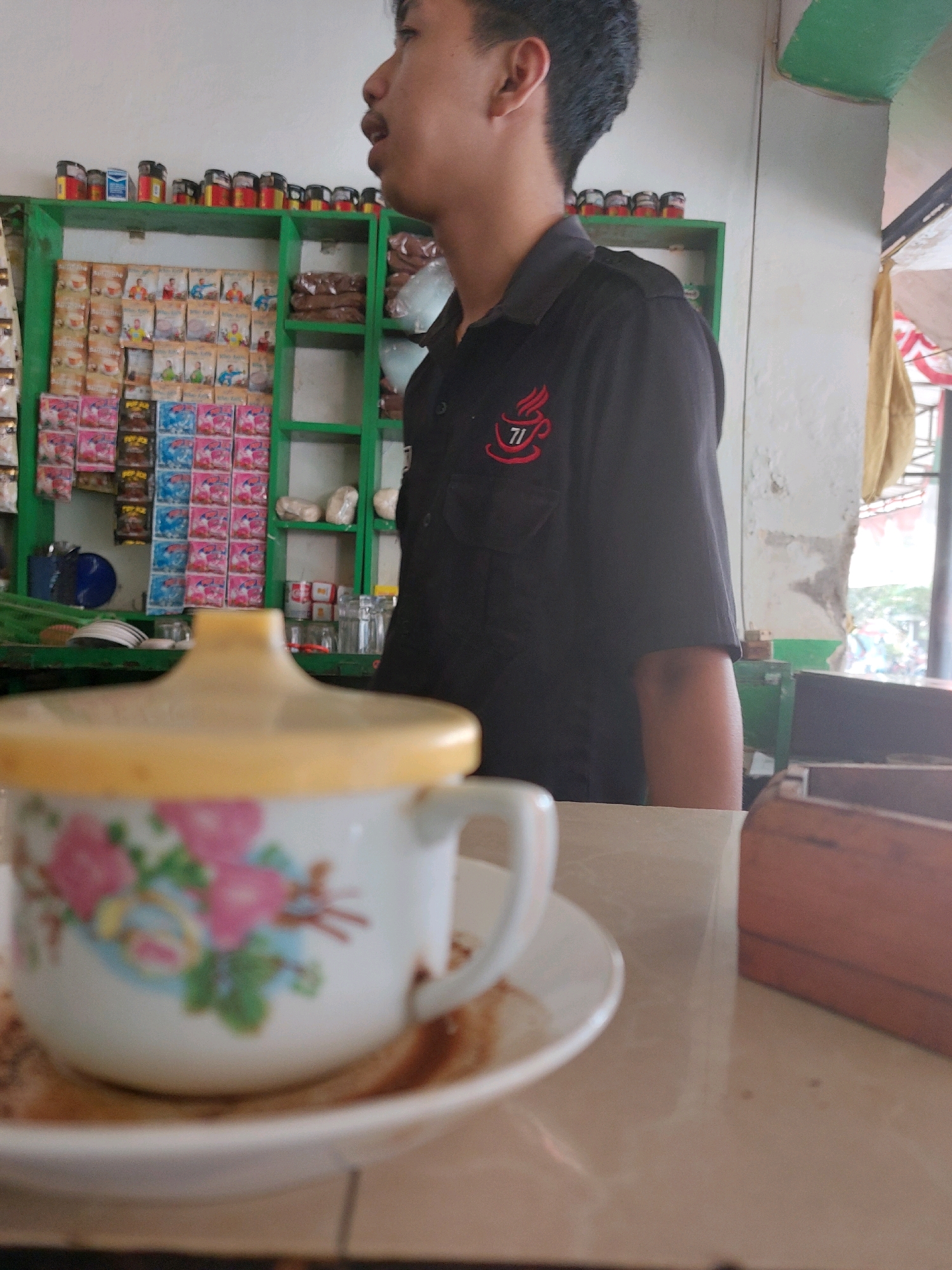 In Sumuragung, Indonesia, a young man wearing a black polo shirt with a red coffee cup logo stands in a shop. The shop's shelves are stocked with various packaged goods, including what appear to be sachets of coffee or instant beverages. In the foreground, a floral-patterned cup with a yellow lid rests on a saucer, positioned on a marble-topped table. The scene suggests a retail environment, possibly a small convenience store or warung.
