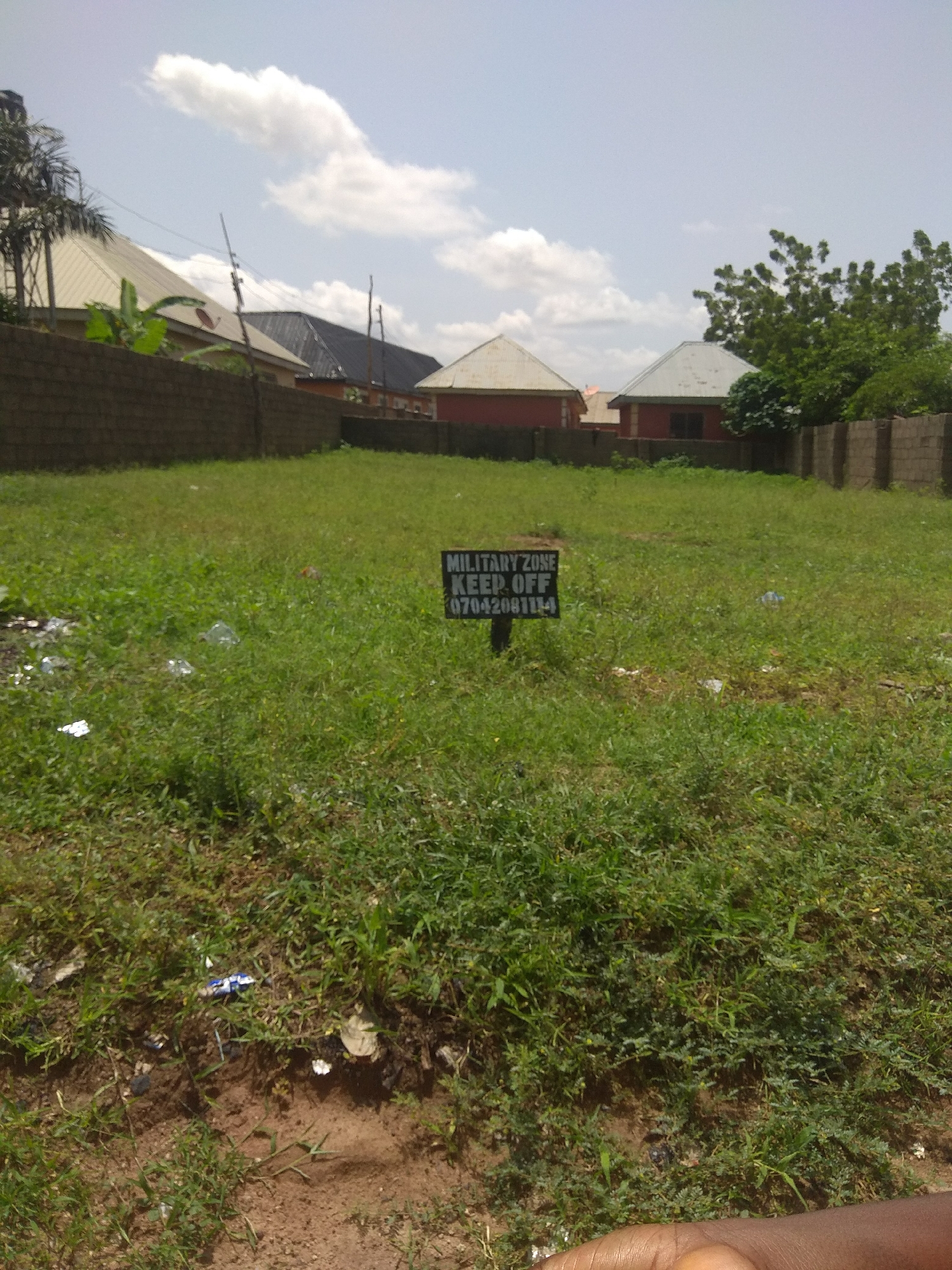 The image depicts an outdoor scene in Jalingo, Nigeria, featuring a vacant lot overgrown with grass. In the foreground, a black signpost with white lettering is visible, reading "MILITARY ZONE KEEP OFF" followed by a phone number "07042081114". The lot is surrounded by a brick wall on the left and a fenced area on the right, with houses visible in the background. The sky is partly cloudy. The ground is uneven and covered in patchy grass with some scattered debris, suggesting neglect. A portion of a person's arm is visible in the lower right corner of the frame, indicating the presence of at least one individual.