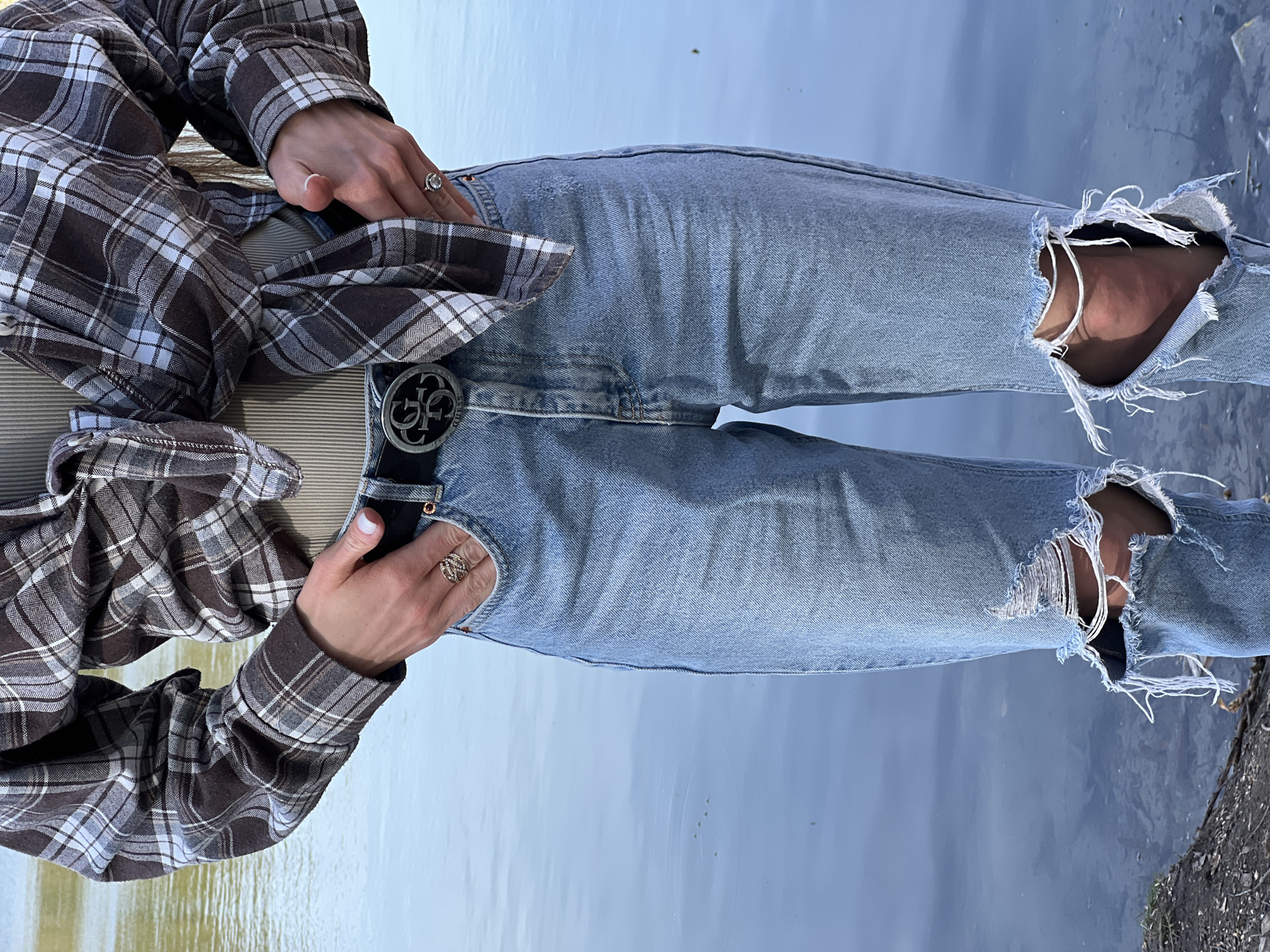 A woman is wearing blue ripped jeans with a large silver "G" belt buckle and a brown plaid shirt in an outdoor setting in what appears to be Ciemne, Poland. Her hands are in the jean pockets, and she is wearing rings. The background is a blurry body of water and some foliage.