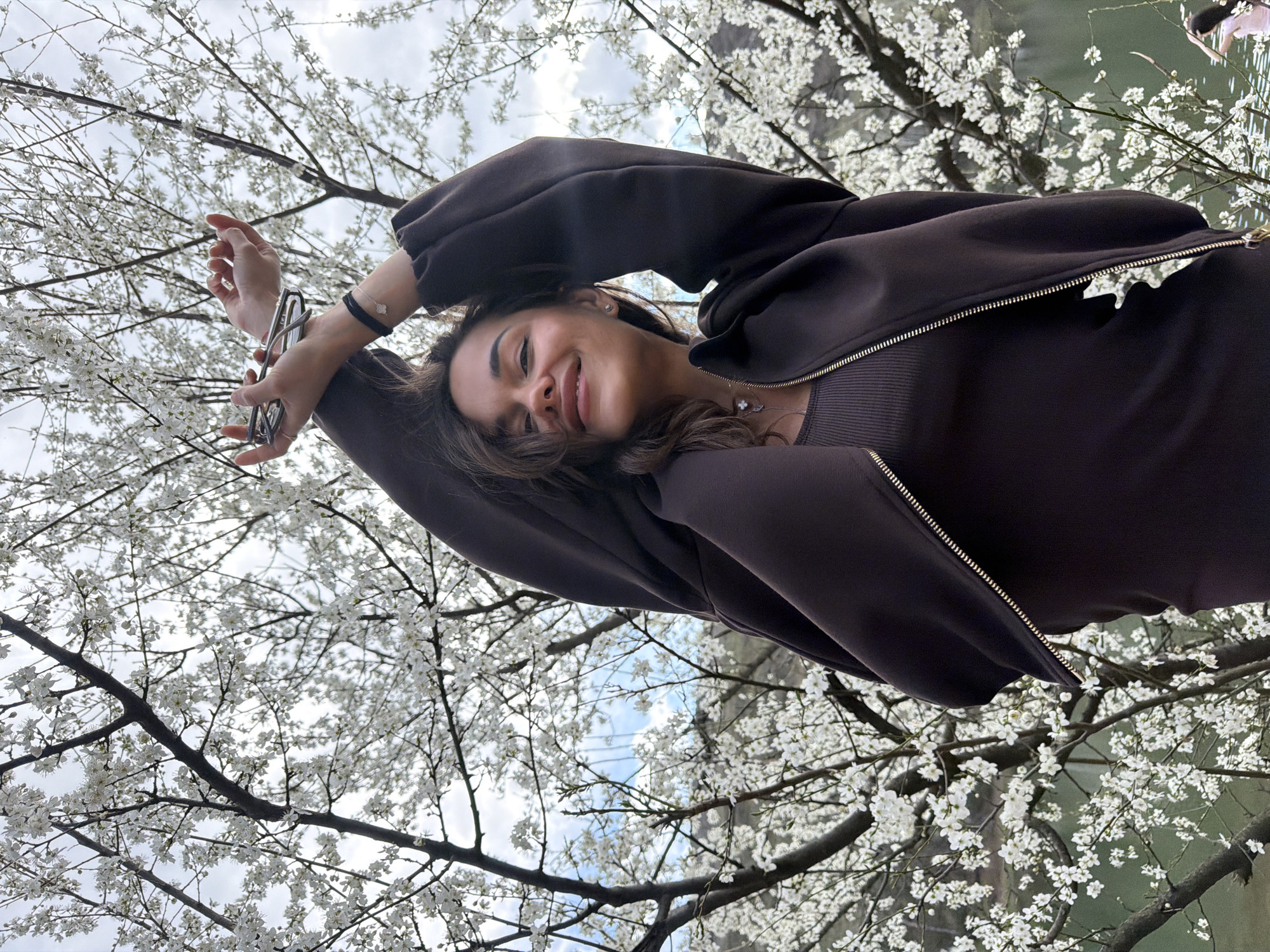 The image shows a young woman from a low-angle perspective, looking up towards her, positioned against the backdrop of a blossoming tree and a body of water. She is wearing a dark brown, possibly ribbed, top with a matching open zip-up jacket. She is holding glasses in her left hand. The background features white blossoms covering the tree branches. There is also a glimpse of what appears to be a lake or river in the background. The location appears to be within the city of Tbilisi, Georgia based on the landscape and architecture that is specific to Georgia.