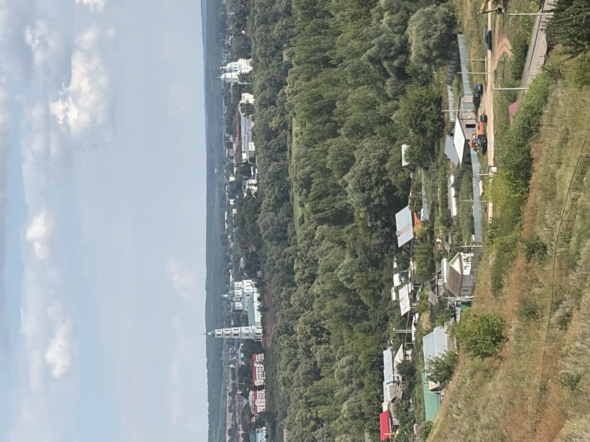 This is an elevated, wide shot of the city of Yelabuga, Russia, on a bright, partly cloudy day. The foreground consists of a steep, grassy embankment covered in dry, golden grasses and scattered green bushes. A faint wire fence is visible in the lower left corner. Below the embankment, a small village or settlement is nestled amongst trees, featuring houses with red and gray roofs, and some smaller outbuildings. A dirt road winds through the village, with a few cars parked near a brick building with an orange truck.
In the mid-ground, a dense forest of green trees extends across the landscape. Beyond the forest, on a distant hill, a prominent white church complex with green domes and a tall bell tower stands out against the horizon. Further in the distance, other buildings and structures of the city are visible, some with light-colored facades and tiled roofs. The sky above is a pale blue, with scattered white cumulus clouds.