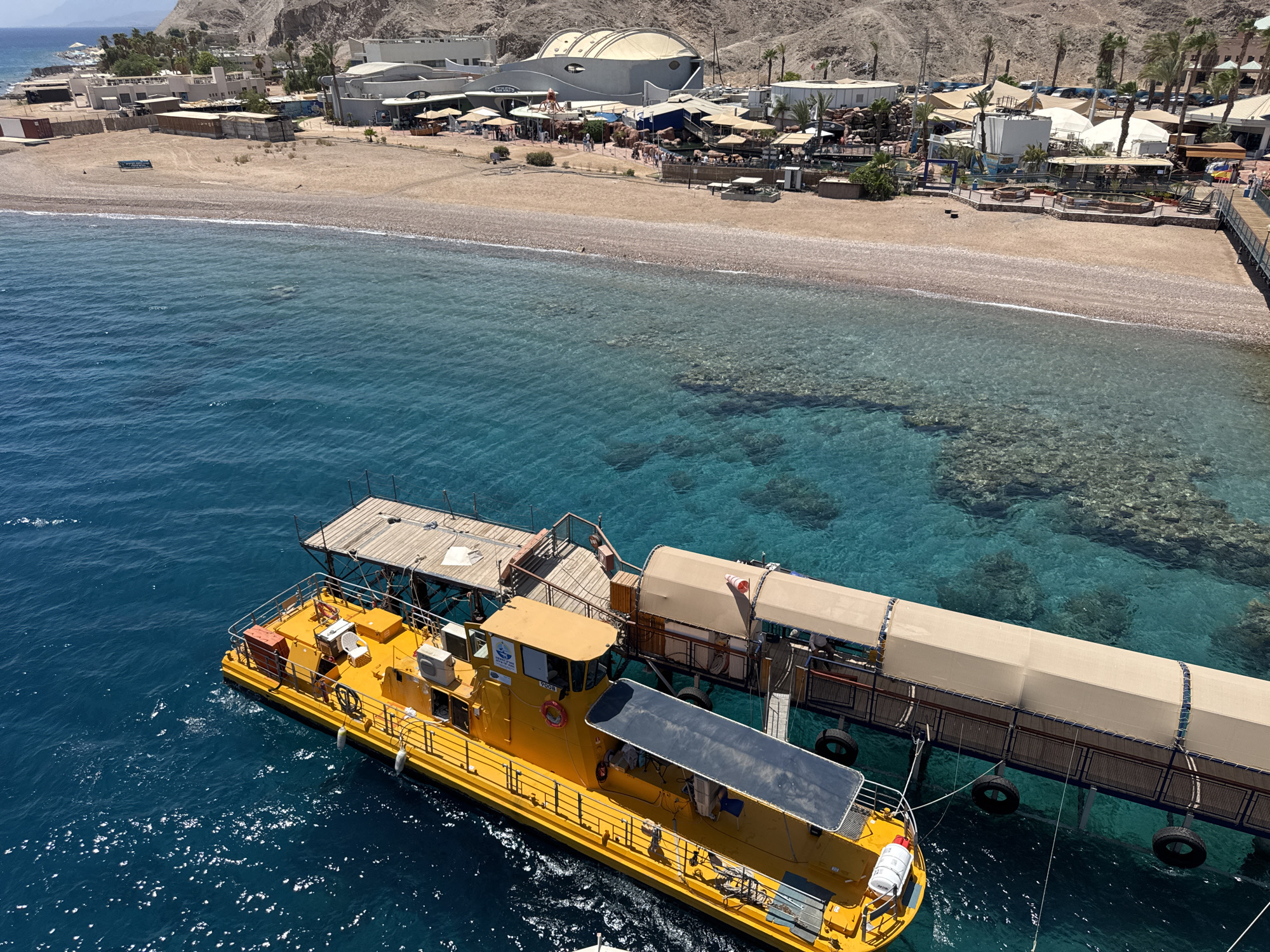 This aerial photograph captures a scene in Eilat, Israel, likely near a coral beach. In the foreground, a bright yellow boat with a covered section and various equipment is docked next to a wooden pier. The boat is moored to a longer covered structure, also accessible by a walkway. The water is a clear turquoise blue, revealing coral reefs beneath the surface.
In the background, a sandy beach stretches along the coastline. Behind the beach, there is a developed area with buildings, shaded seating areas, and palm trees. The architecture suggests a resort or entertainment complex, with some buildings featuring large, modern designs. The landscape behind the developed area is arid and mountainous. Sunlight illuminates the scene, creating sparkling reflections on the water. The overall impression is a sunny day at a coastal tourist destination.