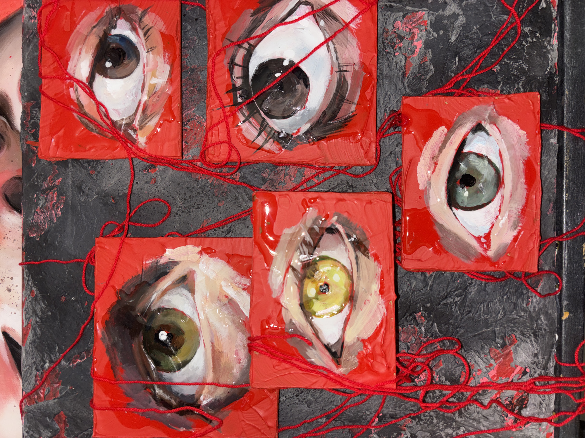 The image displays a close-up of a mixed-media artwork featuring five small, square painted blocks, possibly part of a larger installation. Each block showcases a human eye, rendered with visible brushstrokes on a vibrant, glossy red background. The eyes vary in iris color, including blue, dark brown, green, and yellow-green, and appear to be looking in different directions.
These five eye-themed blocks are arranged against a textured, dark background, which is predominantly black or dark grey with splatters and streaks of red and lighter paint, creating a somewhat chaotic or distressed aesthetic. Bright red string or yarn is intricately draped and woven around and between the blocks, forming a visual network that connects them across the dark surface.
On the far left, a partial view of another artwork is visible, depicting a segment of a human face, including a forehead and eye area, rendered in lighter tones. This suggests the image captures a detail of a broader artistic creation.
The artwork creates an intense and intriguing atmosphere. The striking red of the blocks and string contrasts sharply with the dark background, conveying a sense of drama, emotion, or perhaps a theme of connection or entanglement. The collective gaze of the painted eyes evokes feelings of observation or introspection. The setting appears to be an indoor environment, such as an art studio or gallery, consistent with an artistic display originating from or exhibited in Krasnoyarsk, Russia. No visible text is present in the image.