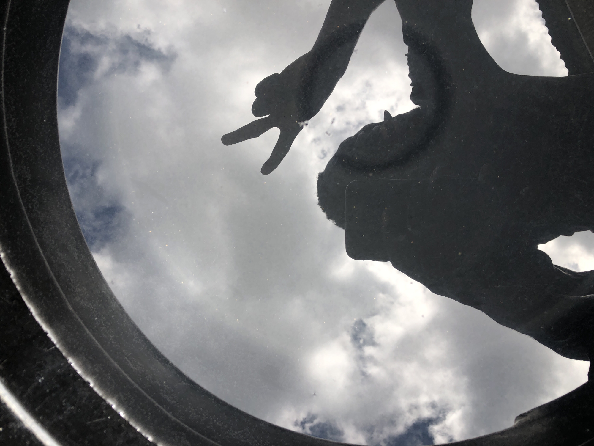 The image, taken from a low-angle perspective, shows a silhouette of a person reflected in a reflective surface, possibly a mirror or a dark, glossy object. The person is making a "rock on" hand gesture, with their index and pinky fingers extended. They appear to be holding a phone, which is also silhouetted. The background of the reflection is a cloudy sky. The outer edge of the reflective surface, seen in the foreground, is a thick, dark, circular rim, possibly part of a tire or a large circular object. The scene is not identifiable as being in the city of Jos, Nigeria, as there are no specific landmarks or context to indicate this location. The image appears to be a self-portrait created by reflecting the sky and the person's gesture in a surface.
