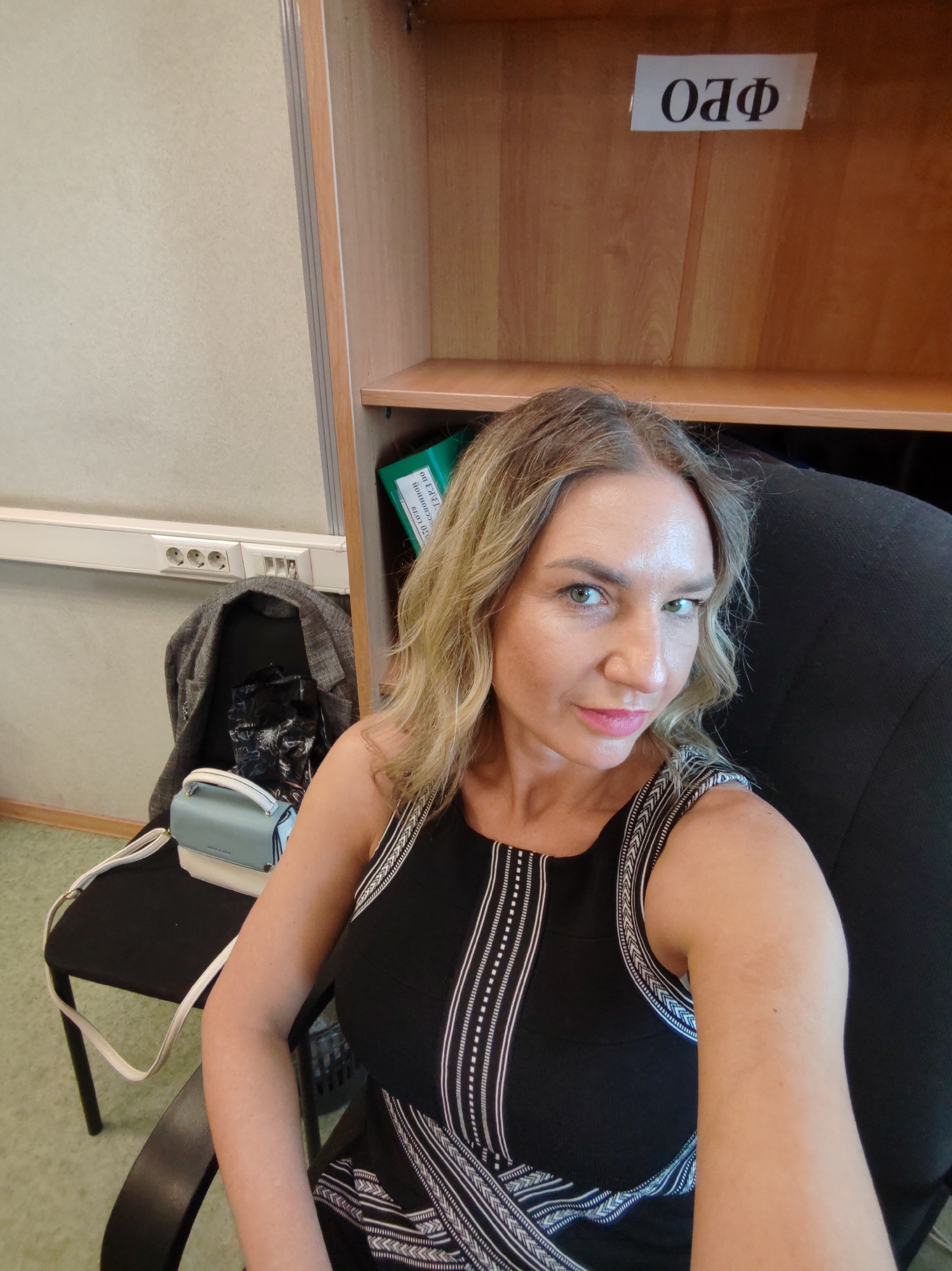The image depicts a woman seated in a black office chair, facing the camera. She has blonde hair and is wearing a black dress with white detailing. Behind her is a wooden cabinet with the Cyrillic letters "ОЛФ" on a white sign affixed to it. To her left, on a small black chair, sits a light blue and white purse and a gray jacket. The room has a light beige wall and green floor tiles. There are electrical outlets and switches visible on the wall behind the chair. The location appears to be indoors, possibly in an office space. The specific city is not identifiable in the image.