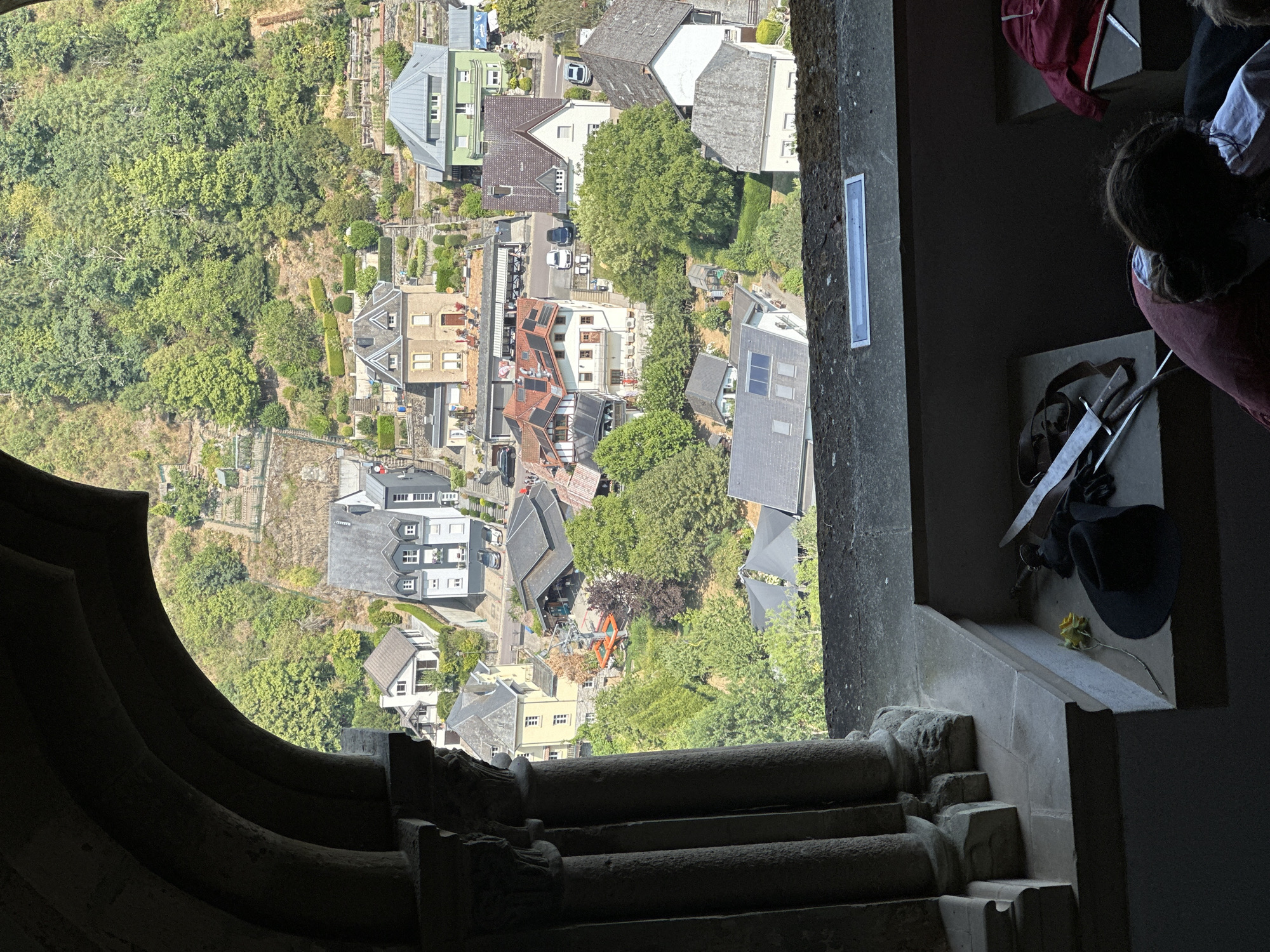 The image captures a view from an arched stone window looking out over the town of Vianden, Luxembourg, with the Vianden Castle possibly in the background obscured by trees. The foreground shows a stone ledge with various objects, including a black hat, gloves, a yellow rose, and two metallic objects resembling swords or large knives. Further back, the town's architecture is visible, characterized by diverse building styles and colors, including houses with red, gray, and green roofs. Some buildings feature solar panels. The town is nestled on a hillside with lush green trees and terraced gardens. In the mid-ground, a cable car with an orange cabin is visible. The overall scene is a detailed snapshot of a historical European town, emphasizing the interplay between man-made structures and the natural landscape.