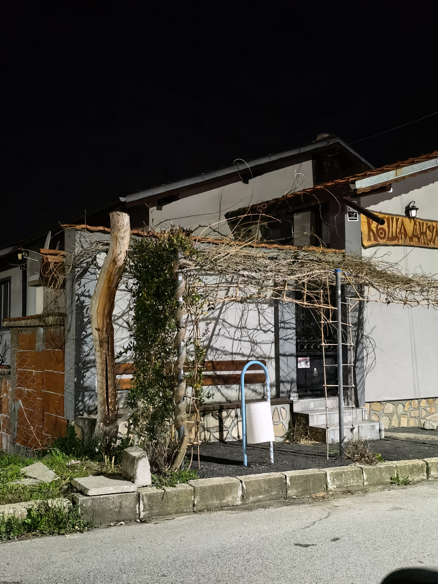 The image shows a building at night. The building has a light-colored facade and a dark roof. A wooden structure covered with vines extends from the building. A bench is visible beneath the structure. A trash can, and a ladder are near the entrance of the building. A sign with what appears to be Cyrillic text is visible above the entrance. The location is city of Unknown, Unknown.