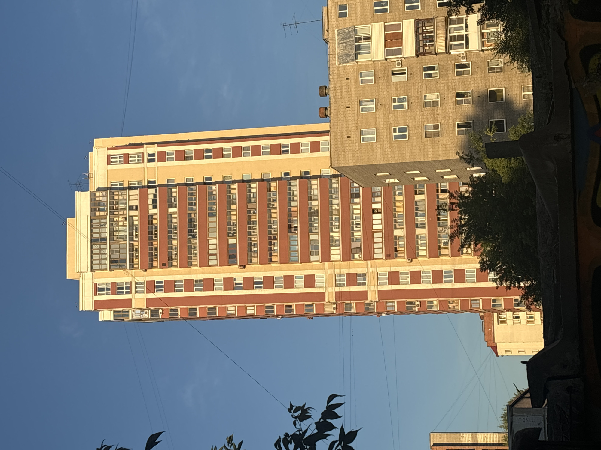 The image captures a view of residential buildings in Novosibirsk, Russia, under a clear blue sky. In the foreground, a tall, slender apartment building with a red and beige facade dominates the center of the frame. The building features numerous windows and balconies, suggesting a high occupancy. To the right of this building, a shorter, wider apartment building with a light grey brick exterior is partially visible. This building also has many windows and a prominent balcony with a glass enclosure. In the background, other residential structures and trees can be seen. Several electrical wires crisscross the sky, typical of an urban environment. The sunlight casts distinct shadows, indicating it is likely late afternoon or early morning. No people or specific activities are discernible in the image. The overall scene depicts a typical residential district in a Russian city.