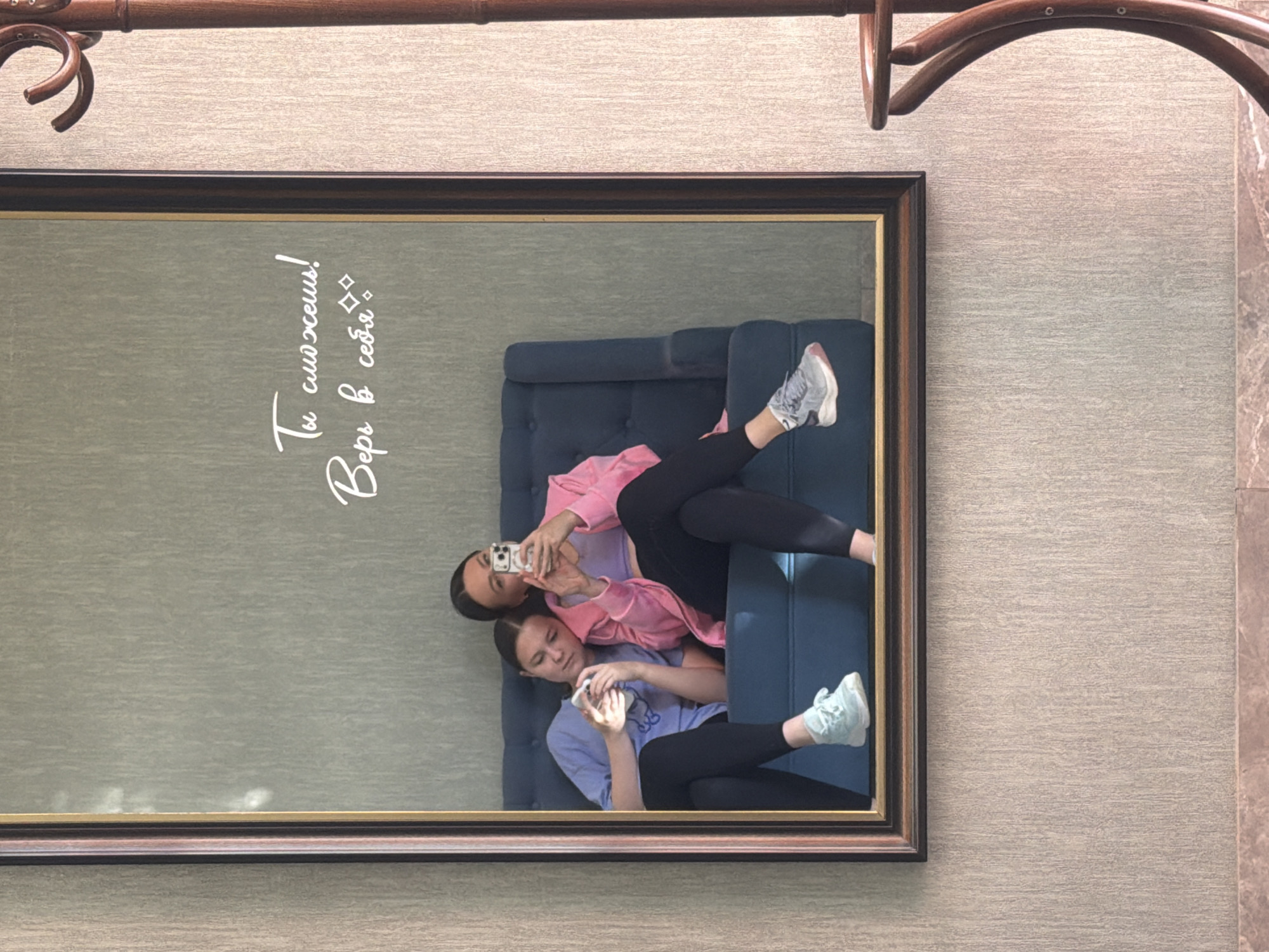A framed mirror reflects two females lying on a blue, tufted couch. One female wears a pink top and black leggings, holding a phone up to take a selfie. The other female wears a light purple shirt and black leggings and is also holding a phone. The mirror hangs on a textured, beige wall. A coat rack is visible at the top of the frame. Text is superimposed on the mirror's reflection. The location, Ovoshchnoy, Russia, is speculative.