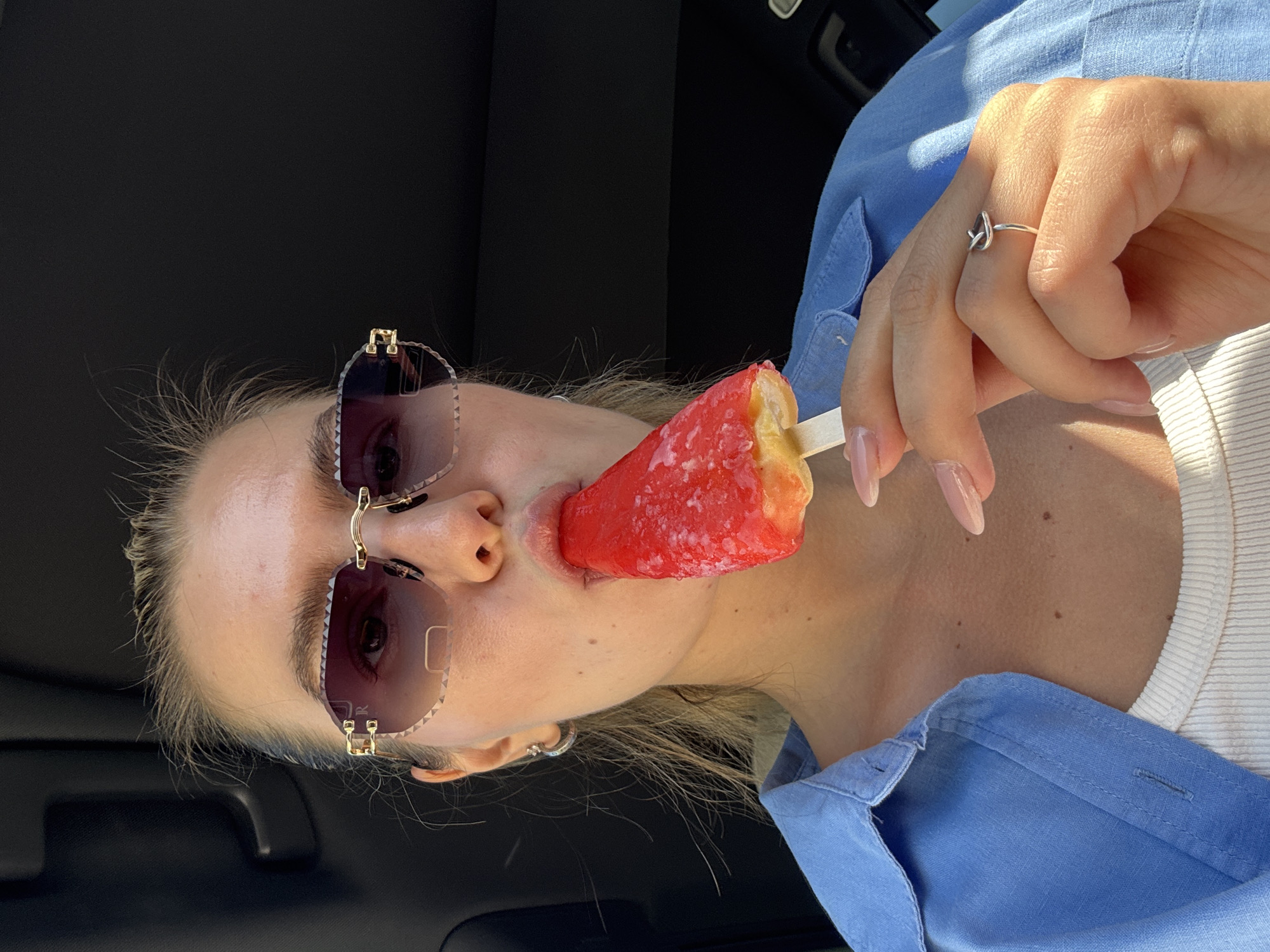 A woman in a blue button-down shirt and white undershirt is seen at an angle, holding a red and yellow popsicle in her mouth. She is wearing gold-rimmed sunglasses and a silver ring. The background is black. The location is unknown.