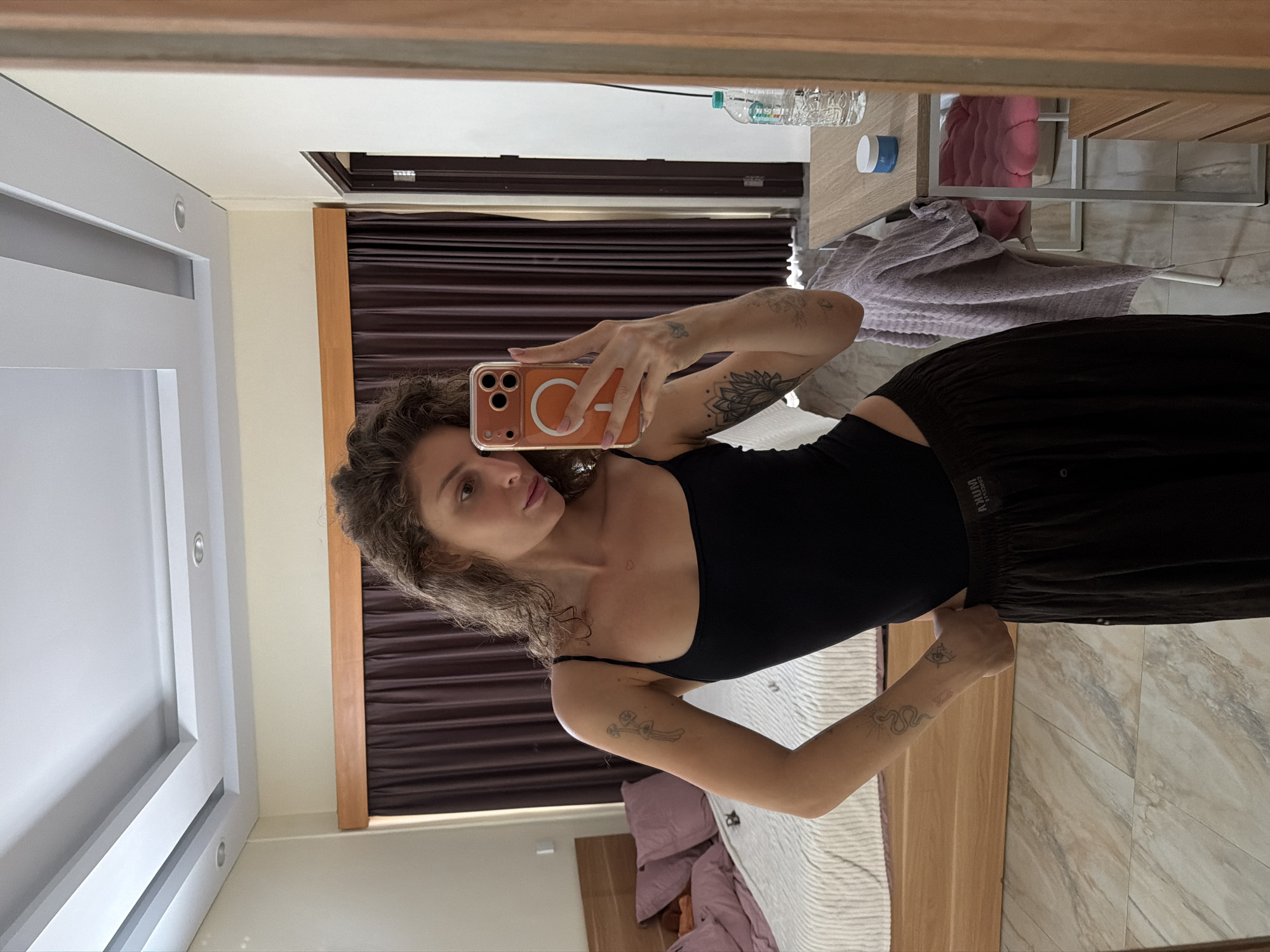 A young woman with curly brown hair, wearing a black tank top and dark brown pants, takes a selfie in a bedroom. She has several tattoos on her arms, including a lotus flower and a snake. The room features a bed with a striped comforter and purple pillows, dark brown curtains, and a wooden dresser. A mirror reflects the woman, a table with a water bottle and a small blue jar, and a closed door. The flooring appears to be light-colored tile. The scene is consistent with a hotel room in North Kuta, Indonesia.