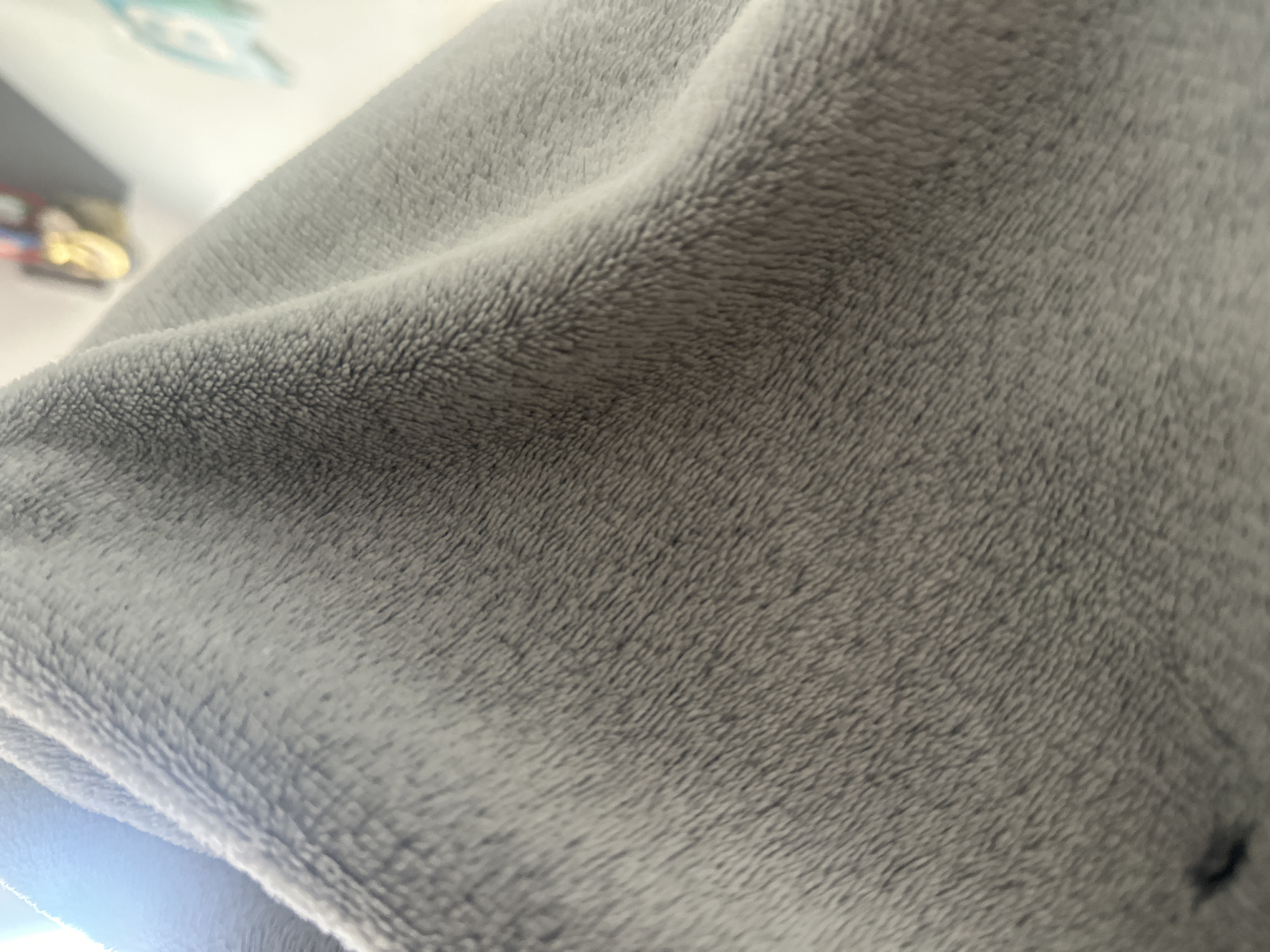 The image displays a close-up, slightly blurred view of a soft, grey, fuzzy fabric, possibly a blanket or a piece of clothing. The fabric exhibits a textured surface with visible fibers. There are subtle shadows and highlights suggesting folds and creases in the material. In the upper left corner, a bright light source is visible, casting a soft glow. In the blurred background, hints of other objects can be perceived, including what appears to be a shelf with items on it, and some green-colored objects. There is a dark, irregular mark on the fabric in the lower left portion of the image. The scene does not provide any visual cues that would allow for the identification of a specific location in the city of Shumen, Bulgaria, or any details about people, actions, or interactions.