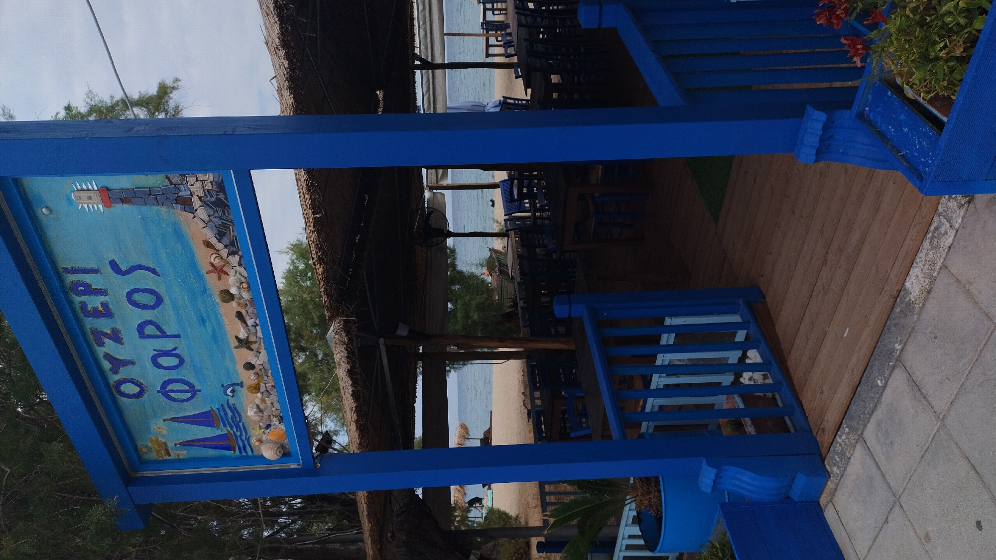 The image shows a restaurant named "OYZEPI FAROS" located in Neoi Epivates, Greece. The restaurant is situated on a sandy beach with the sea visible in the background. The establishment features an outdoor seating area with wooden tables and blue chairs, covered by straw roofs supported by wooden beams. A blue entrance gate with wooden flooring leads into the seating area. A sign above the entrance depicts a lighthouse and maritime elements like sailboats and seashells. The overall ambiance suggests a casual beachside dining experience.
