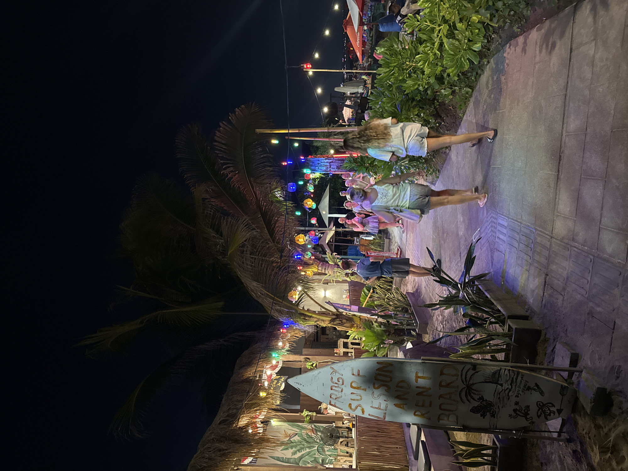 This image depicts a nighttime scene at a beachside establishment in Kuta, Indonesia, likely a surf school and rental shop. A narrow, paved walkway runs alongside a sandy area, bordered by lush green foliage. String lights, including colorful lanterns and red hanging lights, illuminate the scene. On the left, a thatched-roof structure, possibly a bar or shop, is visible with plants and a fan. A prominent surfboard-shaped sign stands in the foreground, displaying text that reads "FROGY SURF LESSON AND RENT BOARD" with accompanying illustrations. Several people are walking along the path, engaged in conversation and enjoying the evening atmosphere. The overall impression is one of a lively, tropical beach town at night.