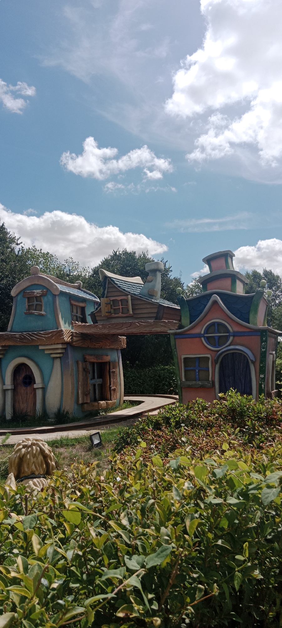 The image shows a whimsical, brightly colored play area with cartoon-like houses and structures set against a backdrop of trees and a blue sky with white clouds. The architecture is exaggerated and playful, with curved roofs, oversized windows, and vibrant colors such as teal, pink, and orange. A pathway winds through the scene. In the foreground, there is dense green foliage and a partial view of a statue or figure, possibly a character from a children's story. The overall impression is one of a cheerful and imaginative outdoor recreational space.
This scene is located within the Parque Europa in Torrejón de Ardoz, Spain. Specifically, it depicts one of the themed play areas within the park, designed to resemble hobbit houses or gnome homes, inspired by fictional dwellings. The park itself is a large green space in the municipality of Torrejón de Ardoz, a city within the Community of Madrid.