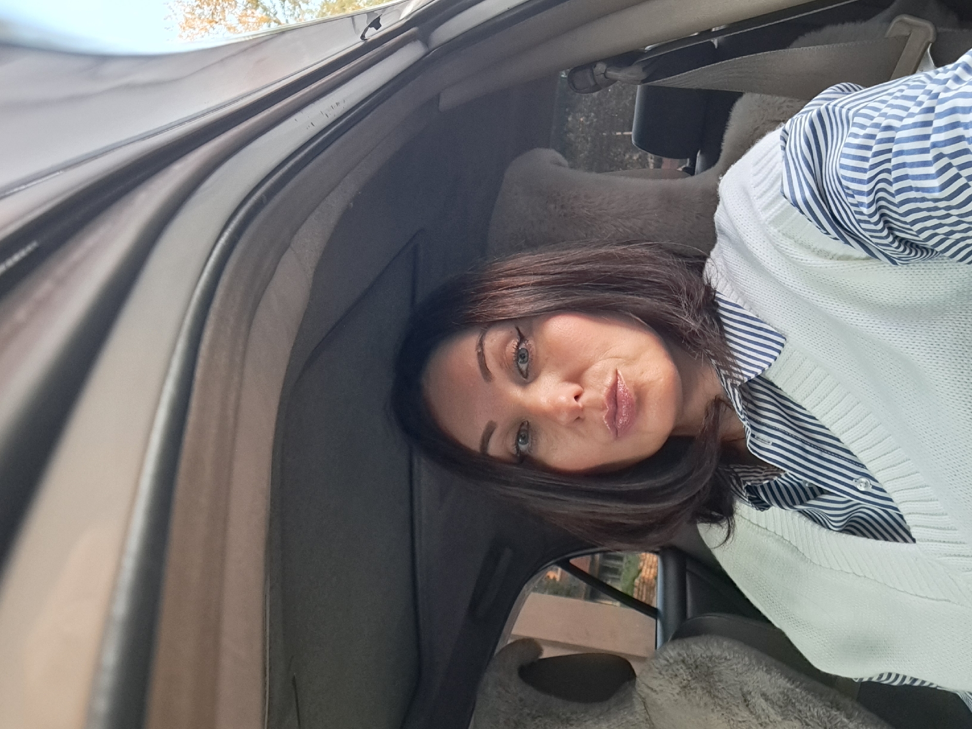 A woman with shoulder-length dark brown hair and light blue eyes is sitting in the driver's seat of a car. She is wearing a white knitted sweater vest over a blue and white striped button-down shirt. Her makeup includes eyeliner and lip gloss. The interior of the car is visible, including the headliner, seats with what appears to be sheepskin covers, and a seatbelt. Through the car windows, trees and a building can be seen, suggesting an outdoor setting. The image is a selfie, taken from a slightly high angle, with the woman looking directly at the camera. The scene does not provide any specific clues to pinpoint the location within Irkutsk, Russia.