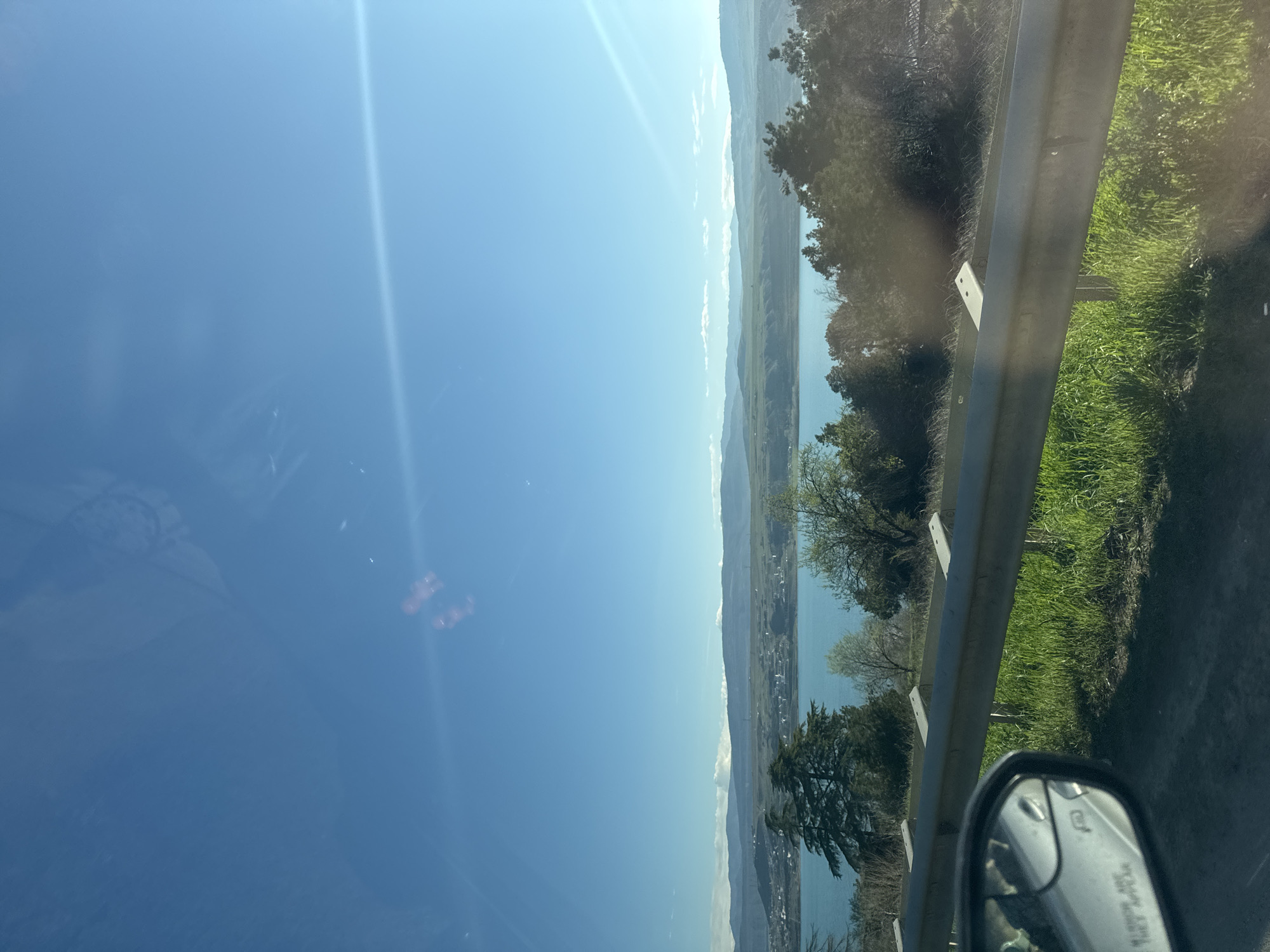 A landscape scene observed from a vehicle, possibly moving on a highway. A body of water is visible with land on the opposite side. Part of a guardrail is in the foreground on the right, along with grass and some vegetation. A side view mirror is visible in the lower right. The sky is clear and blue. It's impossible to pinpoint the exact location in Tbilisi, Georgia from this image.
