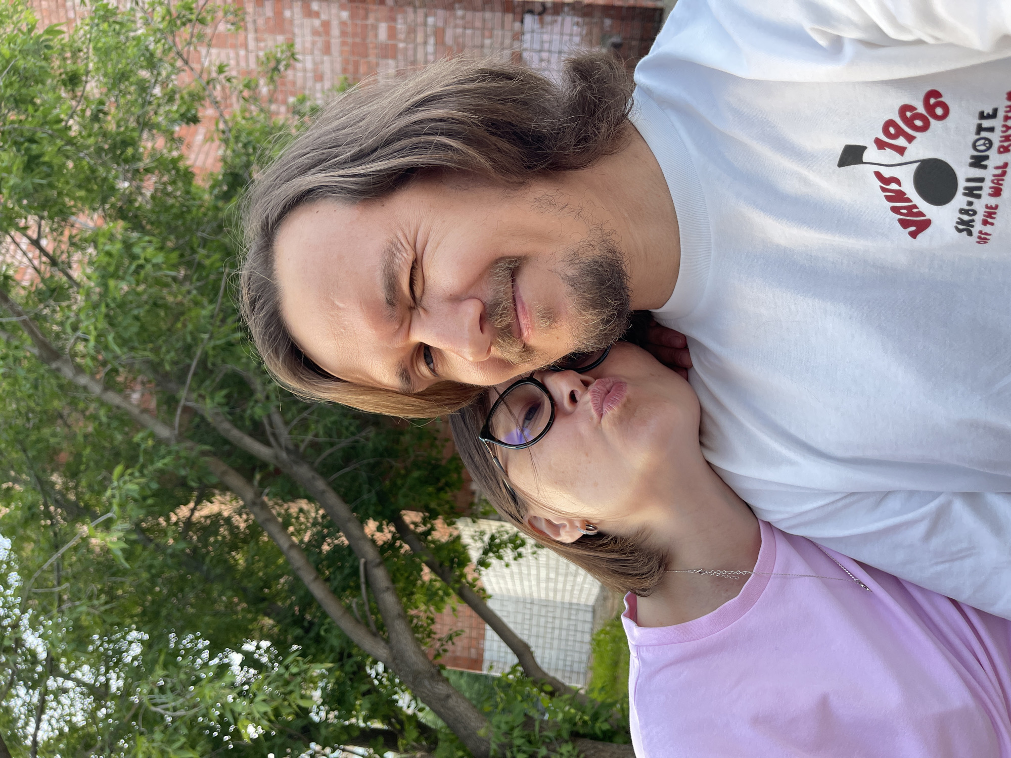 The image shows a man and a woman standing close together outdoors. The man is wearing a white t-shirt with a "Vans 1966" logo on it. He has medium-length brown hair and a beard. He is squinting his eyes. The woman is wearing a pink t-shirt, glasses, and a necklace. She is kissing the man's cheek. A brick wall and green trees are visible in the background. The location may be Izhevsk, Russia, but this cannot be confirmed from the image alone.
