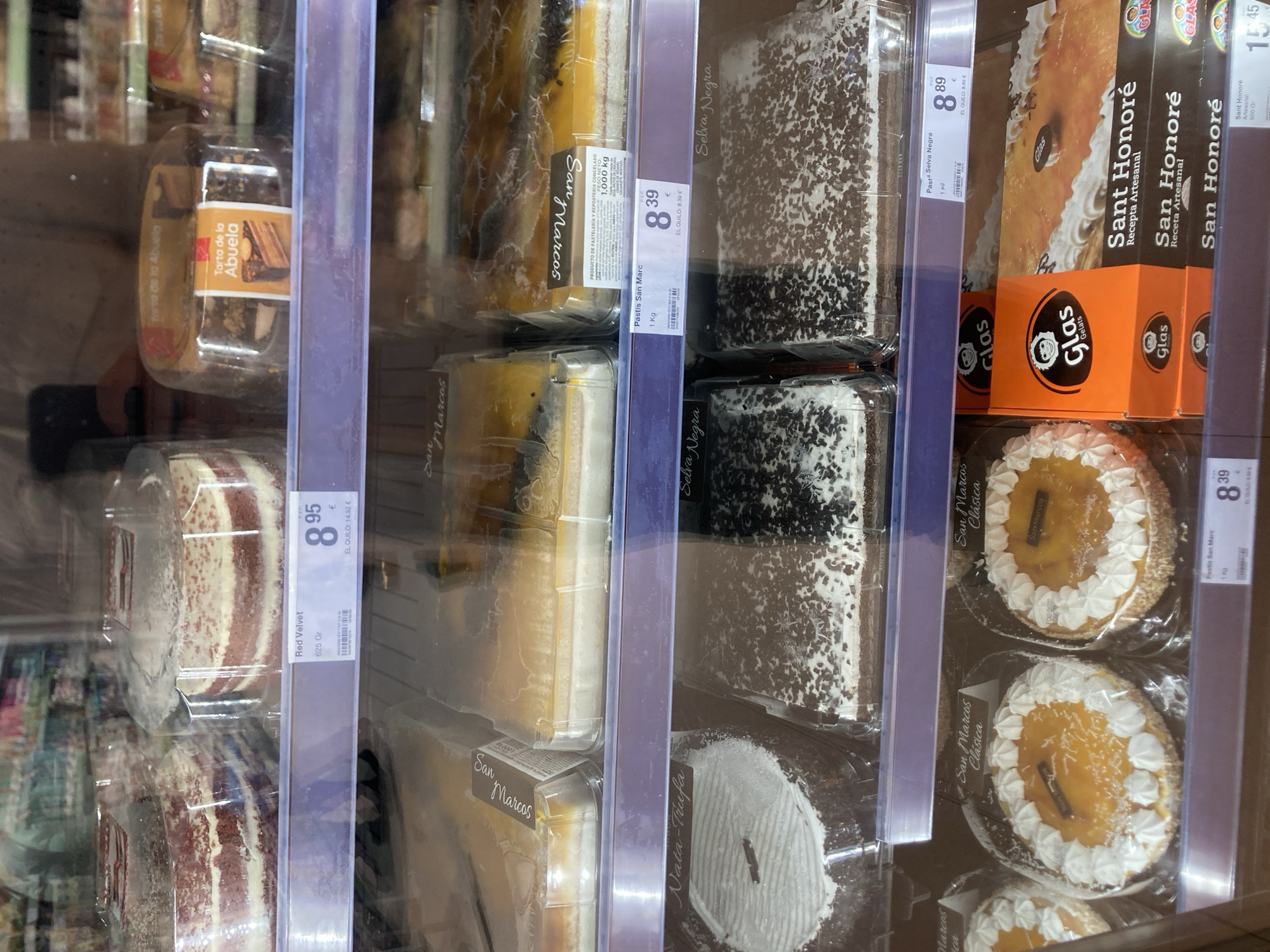 This is a close-up, eye-level, indoor photograph of a refrigerated display case filled with various cakes. The display case appears to be in a supermarket or bakery. The lighting is bright and consistent, suggesting artificial lighting common in such retail environments.
The cakes are packaged in clear plastic containers and are arranged on multiple shelves. The varieties visible include "Red Velvet," "Tarta de la Abuela" (Grandmother's Cake), "Pastís San Marc," and "Selva Negra" (Black Forest). There are also cakes labeled "Sant Honoré" and "Glas Gelats." Some cakes are whole, while others appear to be slices. Price tags are visible for several items, with prices such as 8.95€, 8.39€, and 8.89€. Text on labels indicates weight in kilograms and grams, and prices per kilogram.
The overall scene is clean and organized, typical of a retail setting. There are no people visible in the frame, and the focus is entirely on the products. The location cues, such as the Spanish text on the labels ("Tarta," "Pastís," "Gelats," "Recepta Artesanal," and place names like "San Marcos") and the Euro currency symbols, strongly suggest the setting is in Spain, specifically Barcelona as indicated. The time of day and weather are not discernible from the image. The overall impression is one of abundance and variety in the confectionery offerings.