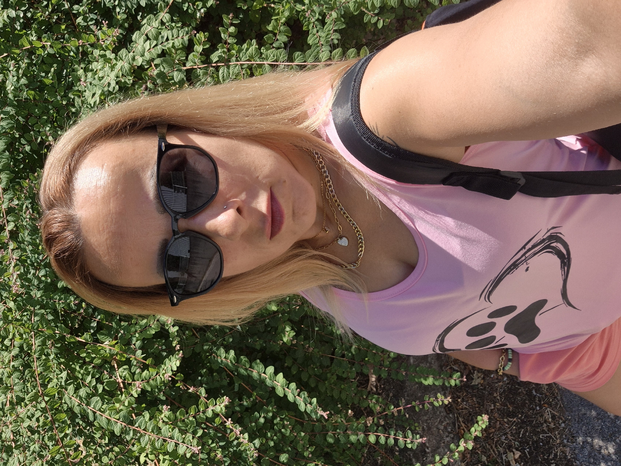 A person with blonde hair is taking a selfie outdoors. They are wearing sunglasses, a pink tank top with a black paw print graphic, and a backpack with black straps. A gold necklace with a small, sparkly heart pendant is visible. They are standing in front of a dense green bush. The ground beneath them appears to be a mix of gravel and dirt.