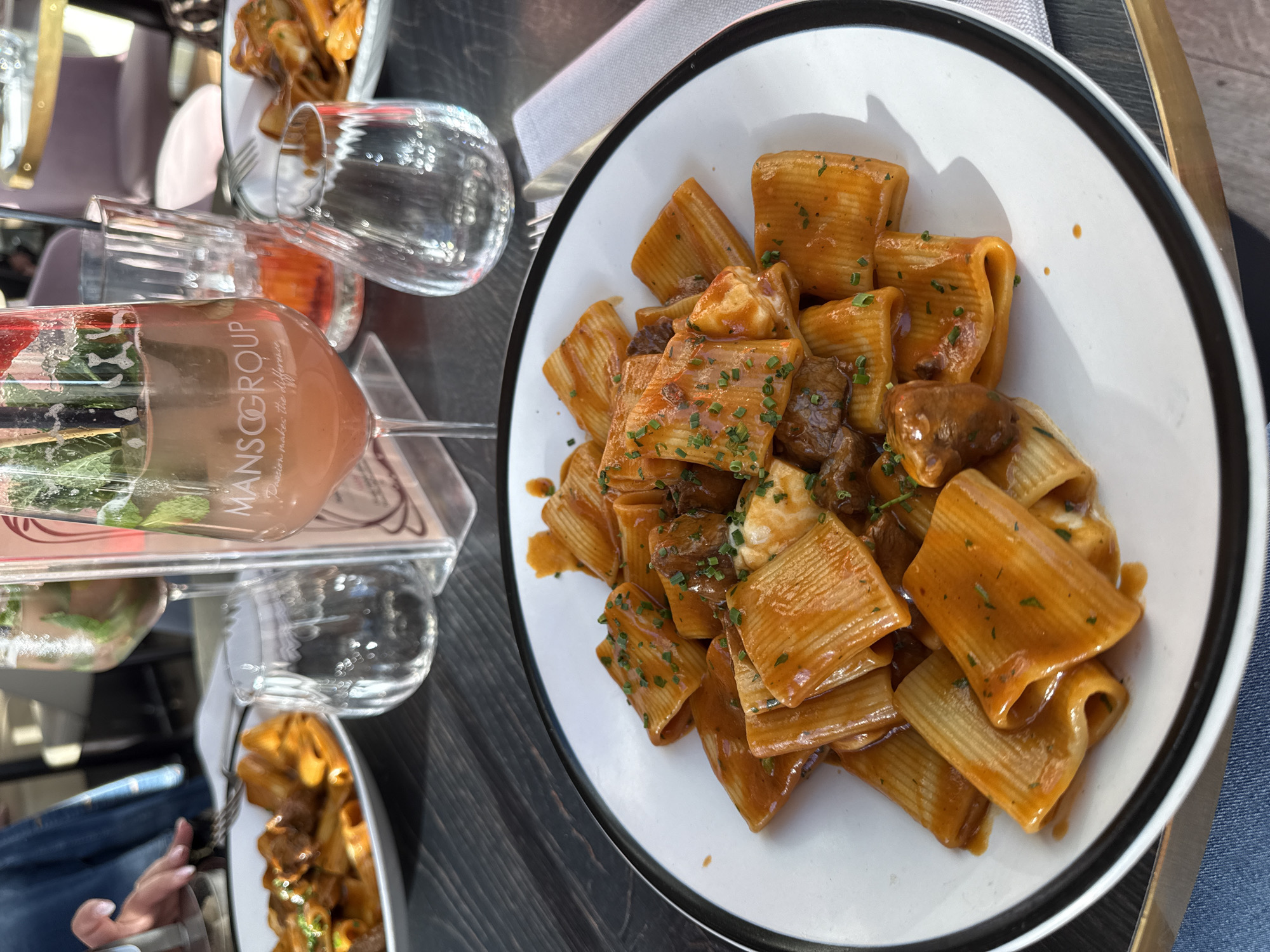 Here is a detailed description of the image:
The image shows a table at what appears to be an outdoor restaurant. A plate of rigatoni pasta with a brown sauce, possibly containing meat and topped with finely chopped herbs, is prominently displayed in the center-right. The plate is white with a black rim.
To the left, there is a half-empty glass of rosé wine labeled "MANSCOGROUP". Next to it, there are two empty glasses. Parts of other plates of the same pasta dish can be seen in the background, suggesting a shared meal.
A person's leg, dressed in blue jeans, is partially visible in the lower right corner. The table surface is a dark gray color. The overall lighting suggests it is daytime, possibly in the late afternoon or early evening. The location is likely Luxembourg City, Luxembourg, as "MANSCOGROUP" is located in that city.