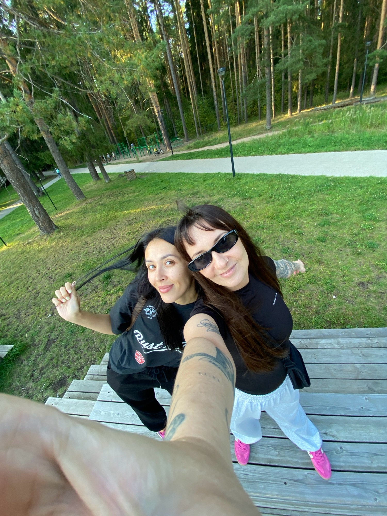 Two women are taking a selfie on a wooden deck in a park in Votkinsk, Russia. The woman on the left has dark hair pulled back and is wearing a black t-shirt and black pants. She is holding a lock of her hair. The woman on the right has dark hair and is wearing sunglasses, a black t-shirt, and white pants. She has tattoos on her arm and is pointing to the right. They are both wearing pink sneakers. In the background, there are trees, a walking path, and a playground. The sun is shining and it appears to be a pleasant day.
