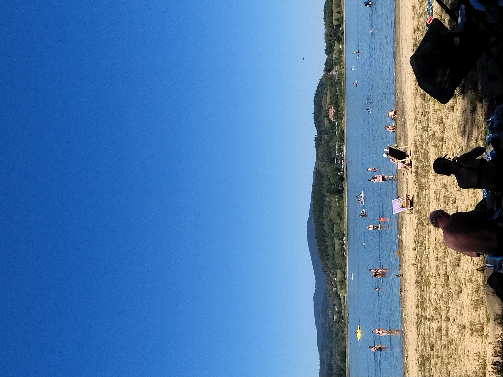 This image depicts a sunny day at a lake with several people engaged in various summer activities. The foreground is a sandy beach with a few individuals seated, two of whom are facing away from the camera, one with a laptop and the other with a book. In the middle ground, a body of water is populated with swimmers, some wading and others further out. Several small boats and watercraft are also visible on the lake. In the background, a lush green, tree-covered hillside with scattered buildings forms the horizon, under a clear, bright blue sky. This scene is located at the Radslav Reservoir, which is part of the recreational area near the village of Radslav, Czechia.