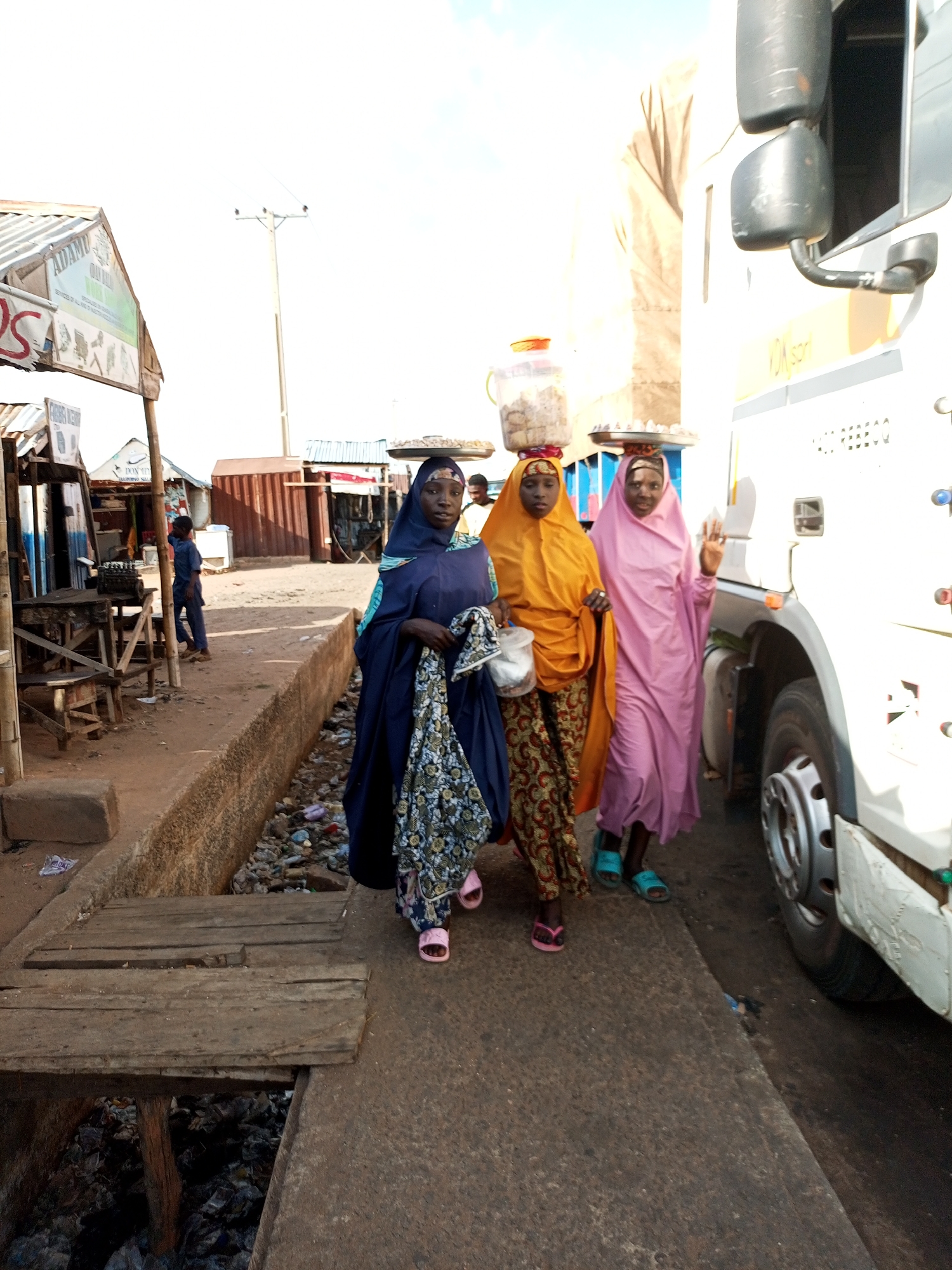 Three women walk along a dirt path next to a large white truck in Gadao, Nigeria. The woman on the left is wearing a dark blue abaya and a patterned scarf, and she is carrying a rolled-up patterned fabric. The middle woman is wearing an orange hijab and a patterned skirt, and she has a round container filled with food balanced on her head. The woman on the right wears a light pink hijab and a matching dress, and she is also carrying a tray of food on her head.
The scene appears to be a market or roadside stall area. To the left of the women are several small structures with corrugated metal roofs, suggesting shops or market stalls. One sign is visible with the text "ADAMU" and "OLAH KALI". In the background, a utility pole stands tall. The ground is dusty and uneven, with a drainage ditch filled with debris running alongside the path. Wooden planks form a makeshift bridge over the ditch. The time of day appears to be daytime, with bright sunlight casting shadows. The overall atmosphere is one of daily activity and commerce.