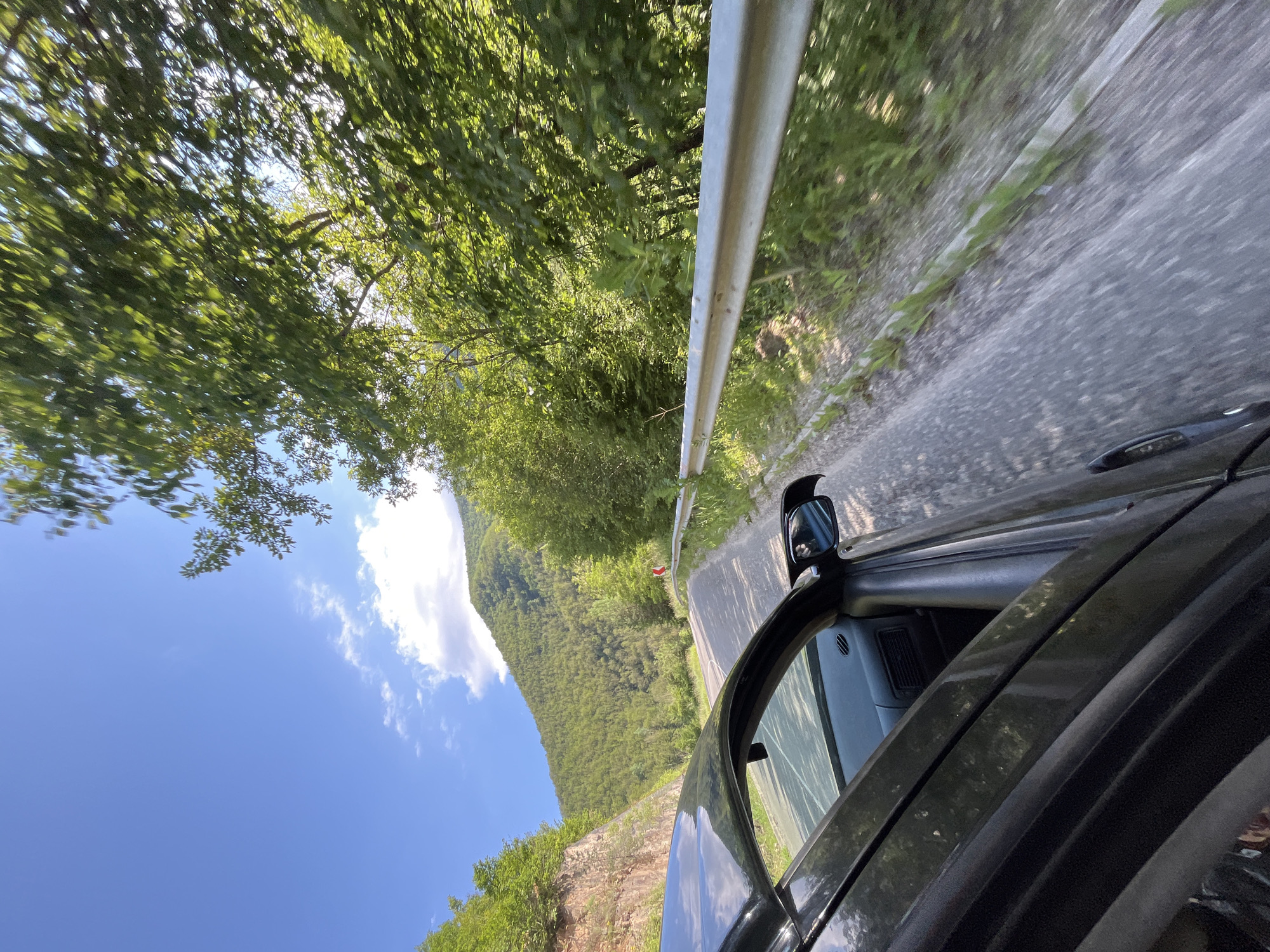 This is a photograph taken from inside a moving car on a winding road in the mountainous terrain near Taran, Bulgaria. The car's dark-colored side and open door are prominently featured in the foreground on the left. The road itself is paved and curves to the right, bordered by a metal guardrail on the right side. Lush green trees and vegetation cover the steep slopes of the mountains that flank the road. The sky is blue with scattered white clouds, and sunlight illuminates the scene. The image conveys a sense of travel through a scenic, natural environment.