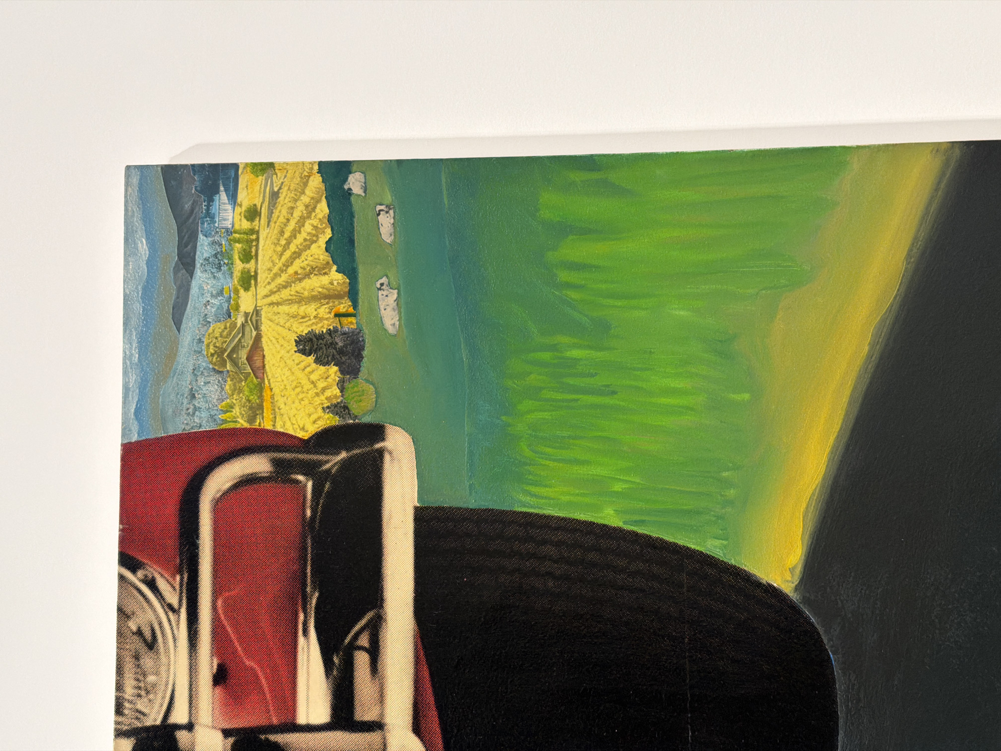The image shows a close-up of a painting on a white wall. The painting depicts a rural landscape with a portion of a red vehicle in the foreground. The landscape features a yellow field with rows, green hills, and distant mountains under a blue sky. Several white shapes, possibly cows, are visible on the green hills. The painting's style appears to be oil on canvas. The artwork is displayed in a minimalist setting, suggesting an art gallery or exhibition space in Tel-Aviv, Israel. There are no people visible in the image.