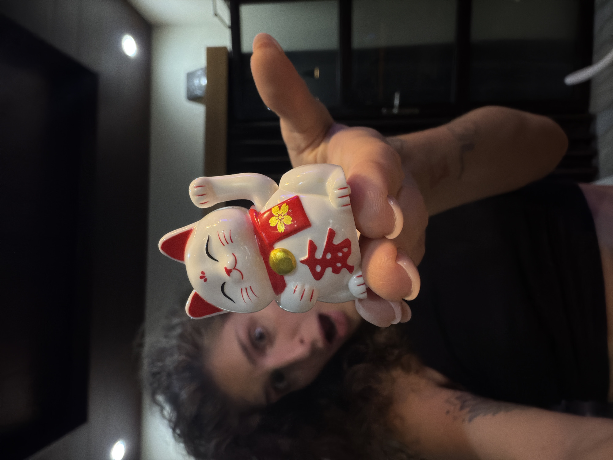 An indoor, close-up shot captures a person holding a Maneki-neko (lucky cat) figurine directly towards the camera. The image is oriented vertically, showing the person upside down with their head at the bottom.
The primary subject is a white Maneki-neko figurine with red accents on its ears and nose. It features closed, smiling eyes and red markings on its cheeks and paws. Around its neck is a red collar adorned with a golden bell and a yellow cherry blossom motif. On its belly, red Japanese characters are visible, appearing to be "福" (fuku), which translates to "good fortune" or "luck." The figurine's left paw is raised, a traditional gesture.
The figurine is held by the fingers of a person, whose hand is prominently displayed behind it. Below the figurine, the person's face is partially visible. They have dark, curly hair, light skin, and several tattoos on their arm. Their expression is one of wide-eyed surprise or playful excitement, with an open mouth. They are wearing a dark-colored top, possibly black.
The setting appears to be a dimly lit interior space, with artificial light sources visible as bright circular reflections on a dark surface in the background. Other blurry background elements include dark and lighter wall sections, and indistinct architectural features. The scene suggests a casual, perhaps playful, moment, possibly a selfie. The location context is North Kuta, Indonesia, though no specific geographical cues are visible within the indoor scene. The lighting suggests it could be evening or night, or an interior not relying on natural light.