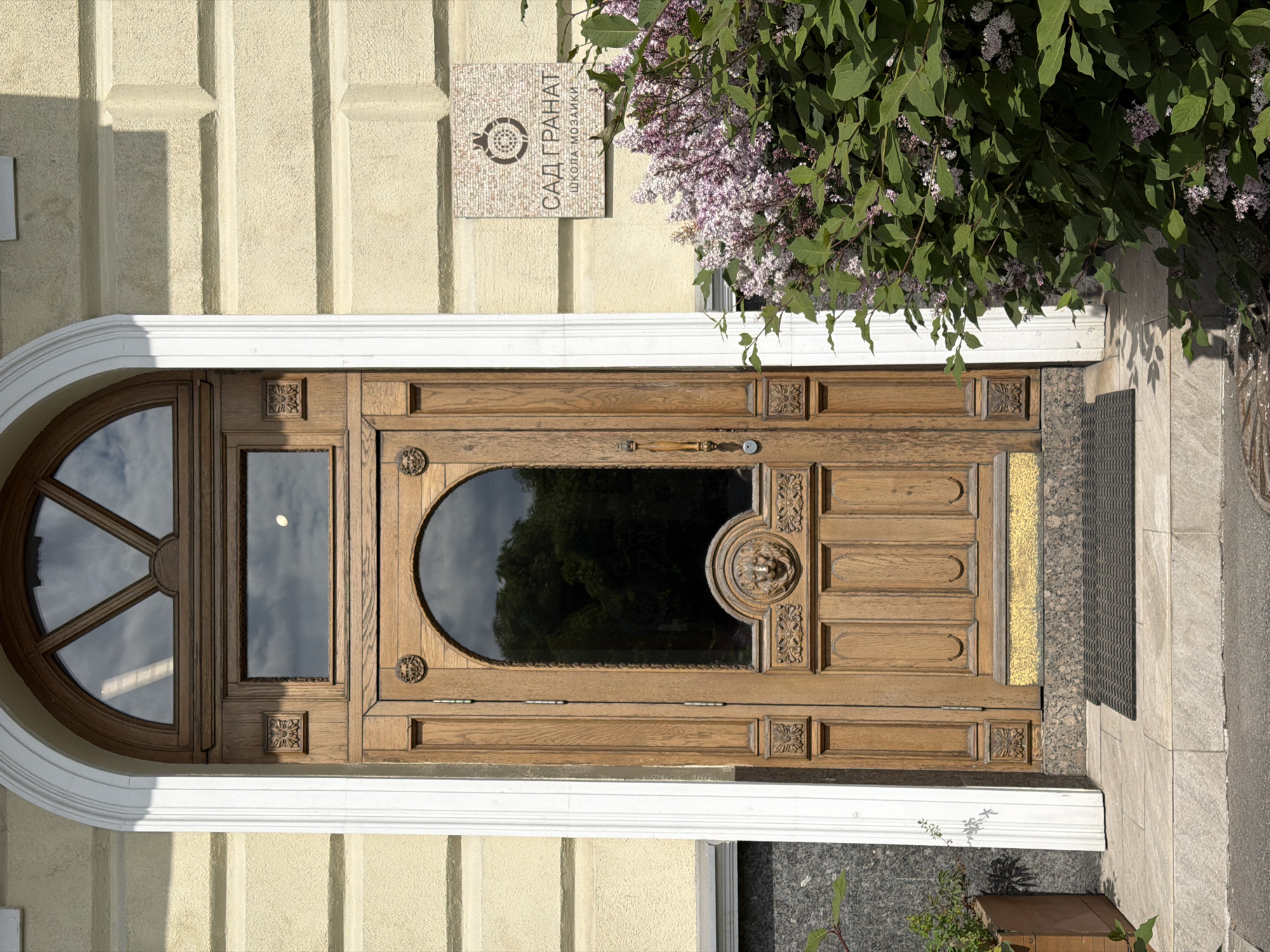 The image displays a brightly lit building entrance in Saint Petersburg, Russia, featuring a richly detailed wooden door. The building's facade is a textured, light yellow-beige with prominent vertical structural elements. A curving white architrave frames the top of the dark brown wooden door.
The door itself is ornately carved with various decorative panels and motifs, including square rosettes and a lion's head relief near the handle. It incorporates several glass panels: a large semi-circular window with radial dividers on the left, a smaller rectangular pane, and a prominent arched glass panel on the right. The glass surfaces reflect the clear blue sky with scattered clouds in the left window and dark green foliage in the larger arched window. A decorative wooden handle is positioned centrally on the door.
Above the entrance, a rectangular mosaic plaque is affixed to the wall. It features a stylized pomegranate icon and Cyrillic text that reads "САД ГРАНАТ" (SAD GRANAT), meaning "Pomegranate Garden," and "ШКОЛА МОЗАИКИ" (SHKOLA MOZAIKI), meaning "Mosaic School."
A lush branch of a lilac bush, adorned with vibrant green leaves and clusters of light purple flowers, hangs gracefully over the upper right section of the door and entrance, casting dappled shadows. The ground directly in front of the door consists of light-colored paving stones, with a dark doormat visible on the right. The scene is illuminated by natural daylight, suggesting clear and sunny weather. No people are visible, emphasizing the architectural and botanical details of the entrance.