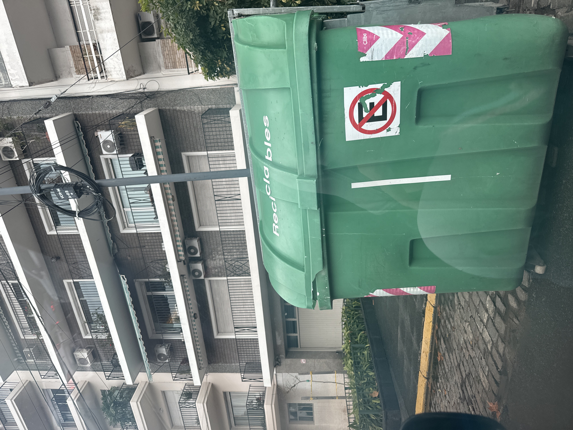 A green recycling bin with "Recicla bles" printed on it is positioned on a sidewalk, adjacent to a yellow curb. The bin has stickers, including one with a red prohibition symbol. Behind the bin, there's a residential building with multiple floors, balconies, and air conditioning units. Utility poles with wires are visible to the left of the building. The location is in Buenos Aires, Argentina based on the Cliba sticker, a waste management company operating in that city. The road and sidewalk appear wet, suggesting recent rain.
