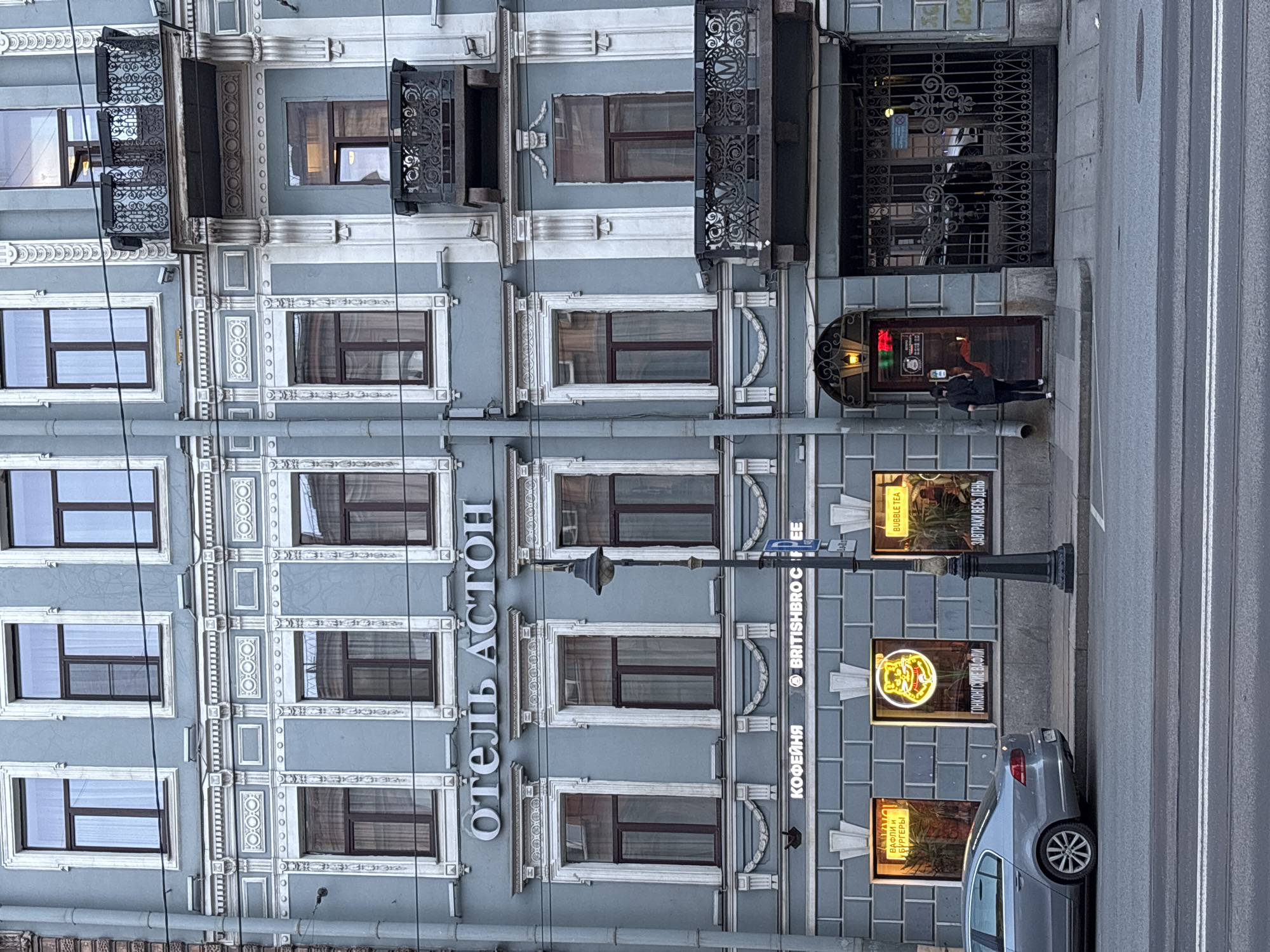 The image shows the facade of a building in St. Petersburg, Russia. On the left side of the building is the vertical text "ОТЕЛЬ АСТОН" (Hotel Aston). To the right of this is a coffee shop called "BRITISHBRO COFFEE". There are three lit signs in the coffee shop windows, advertising "WAFFLES AND BURGERS", "HONG KONG WAFFLES", and "BREAKFAST ALL DAY". In front of the coffee shop is a parked silver car. On the sidewalk is a person in a black jacket and white shoes, standing near the doorway of the building. The architectural style of the building is ornate with decorative window frames and balconies. There is a black iron gate to the right of the doorway. The street is visible in the foreground.