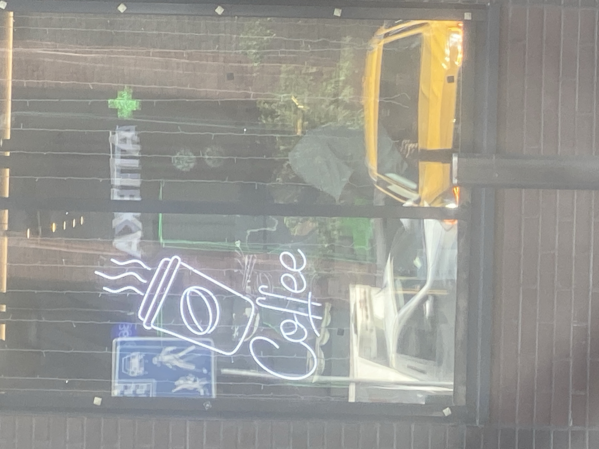 The image depicts a window of a building in the city of Khimki, Russia, showcasing a neon sign that spells out "Coffee" above a stylized coffee cup with steam rising from it. The neon sign is white and illuminated. To the left of the coffee sign, a blue sign with white pictograms is visible, indicating traffic or pedestrian regulations. Through the window, several vehicles are visible, including a white van or truck and a bright yellow car. The yellow car's headlights are illuminated. The background shows greenery and possibly other buildings. The window is framed by vertical strings of lights.