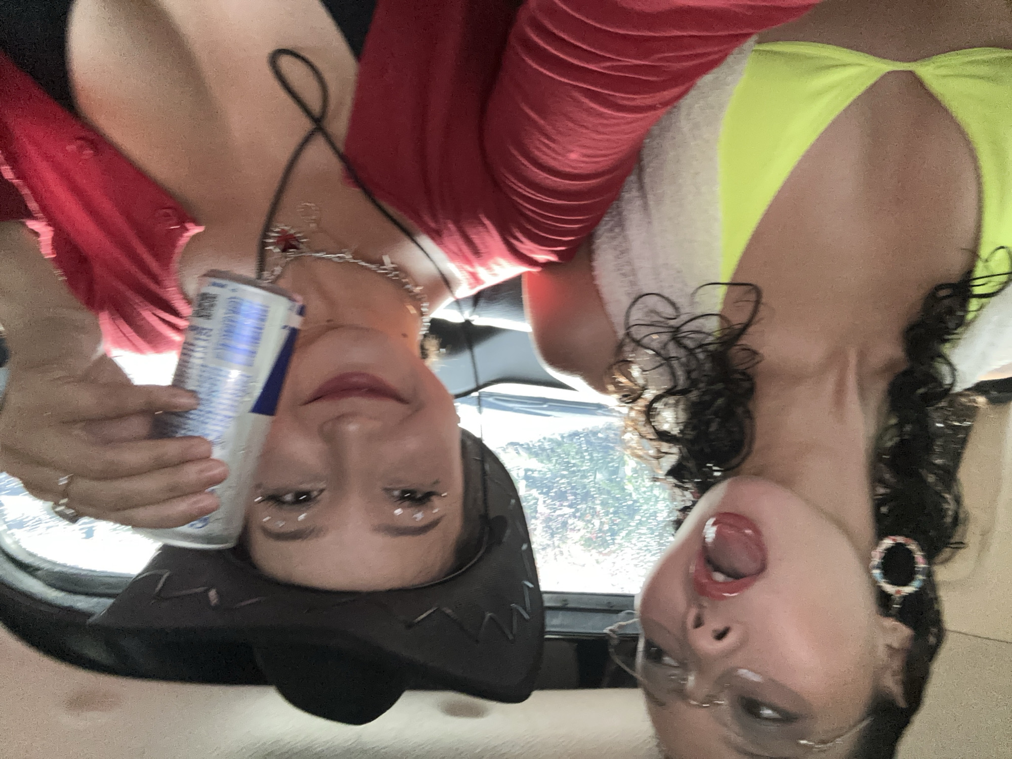Two women are sitting inside a vehicle. The woman on the left wears a black cowboy hat, a red top, and jewelry. She is holding a can of Red Bull. The woman on the right wears a neon yellow top, glasses, and dangling earrings. The background shows greenery, suggesting an outdoor environment. The scene lacks sufficient information to confirm the location of North Kuta, Indonesia.