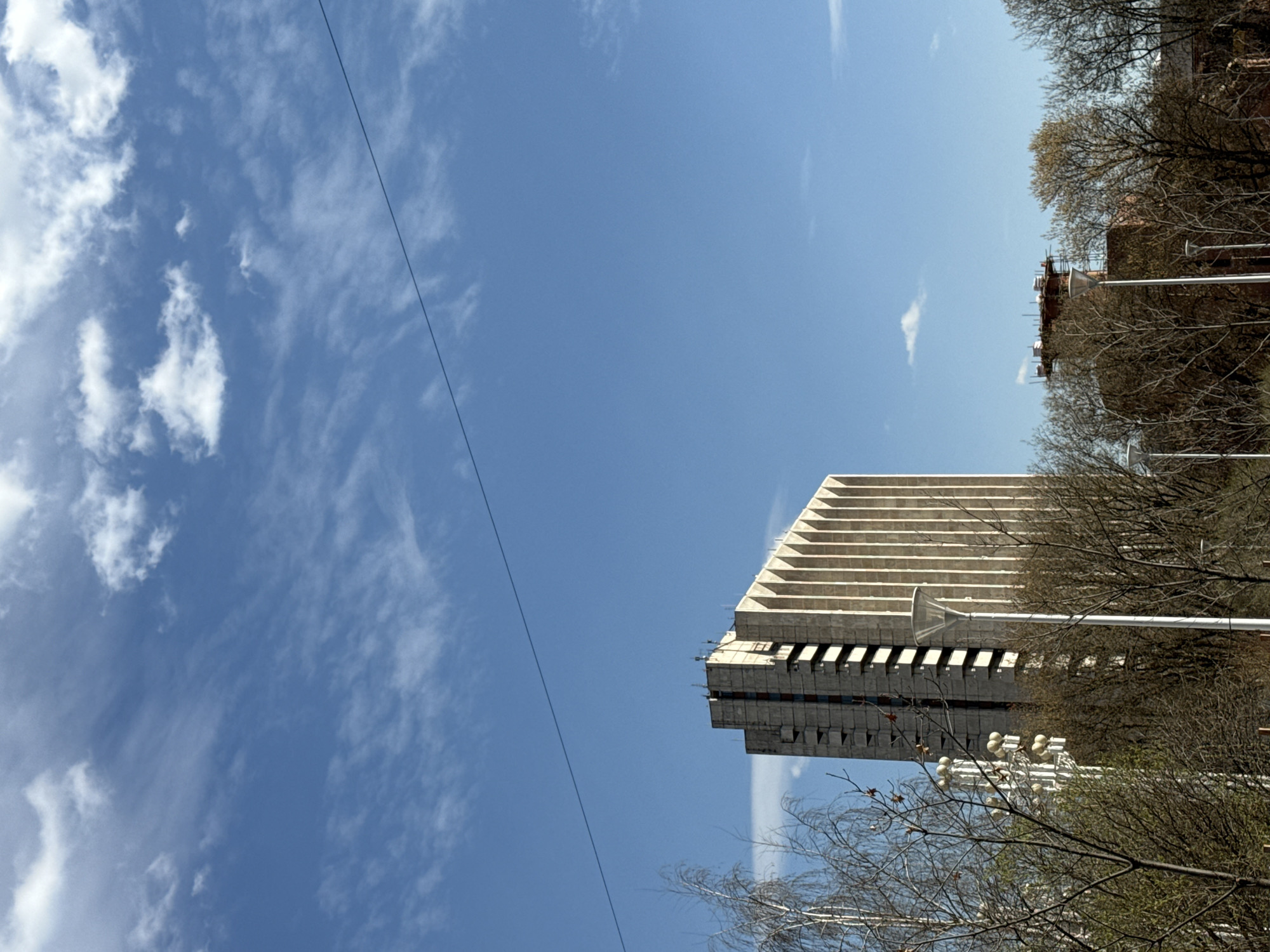 The image shows the State Duma building in Moscow, Russia. The building is viewed from below, revealing a portion of the facade with its distinctive horizontal lines, juxtaposed against a blue sky partially filled with wispy clouds. Bare tree branches frame the right side of the image, partially obscuring the view of the building and adjacent architecture. A power line stretches across the sky from the upper left to the middle of the frame.